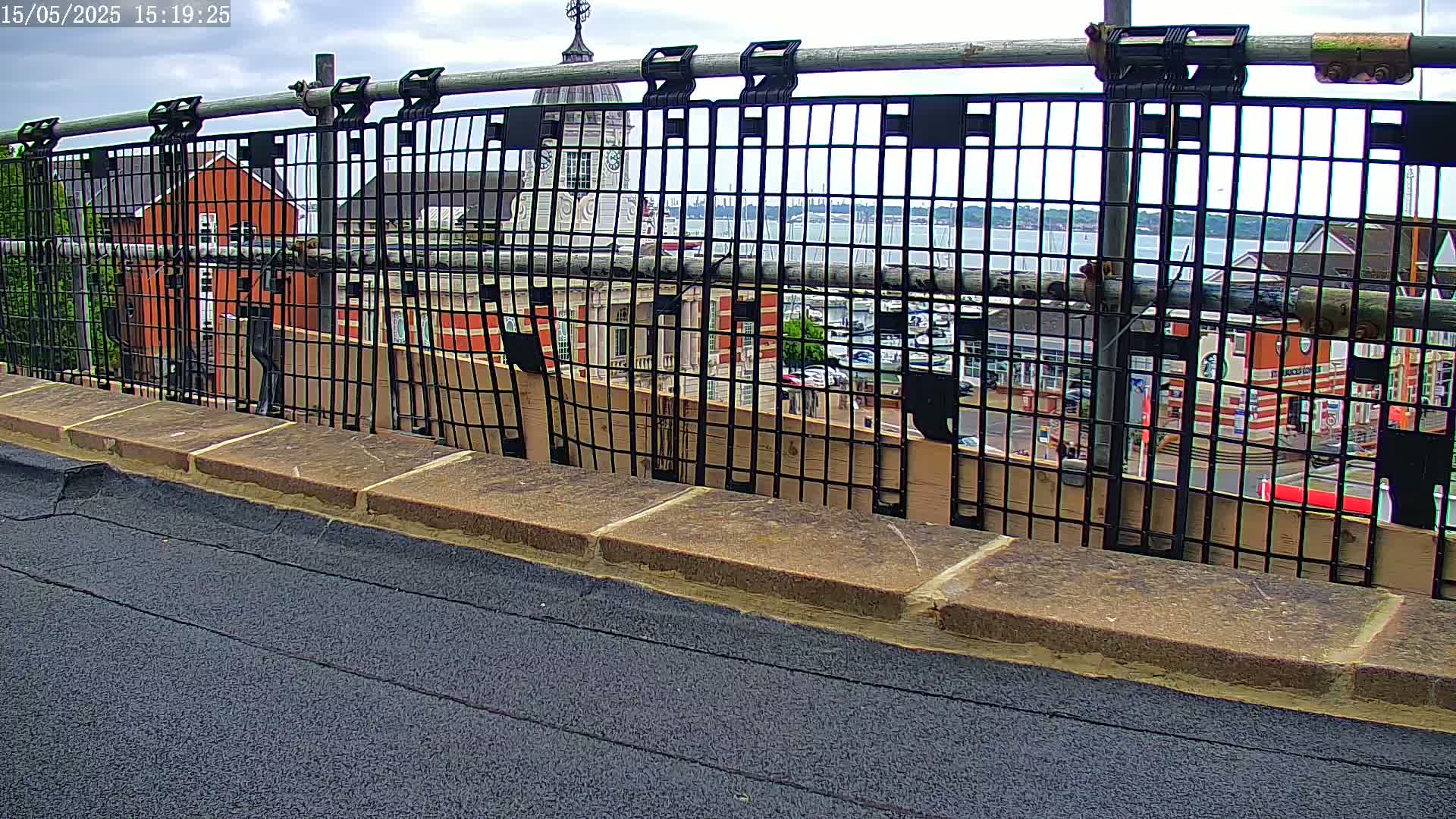 A black metal fence on a rooftop overlooks a town with a body of water and partly cloudy skies.