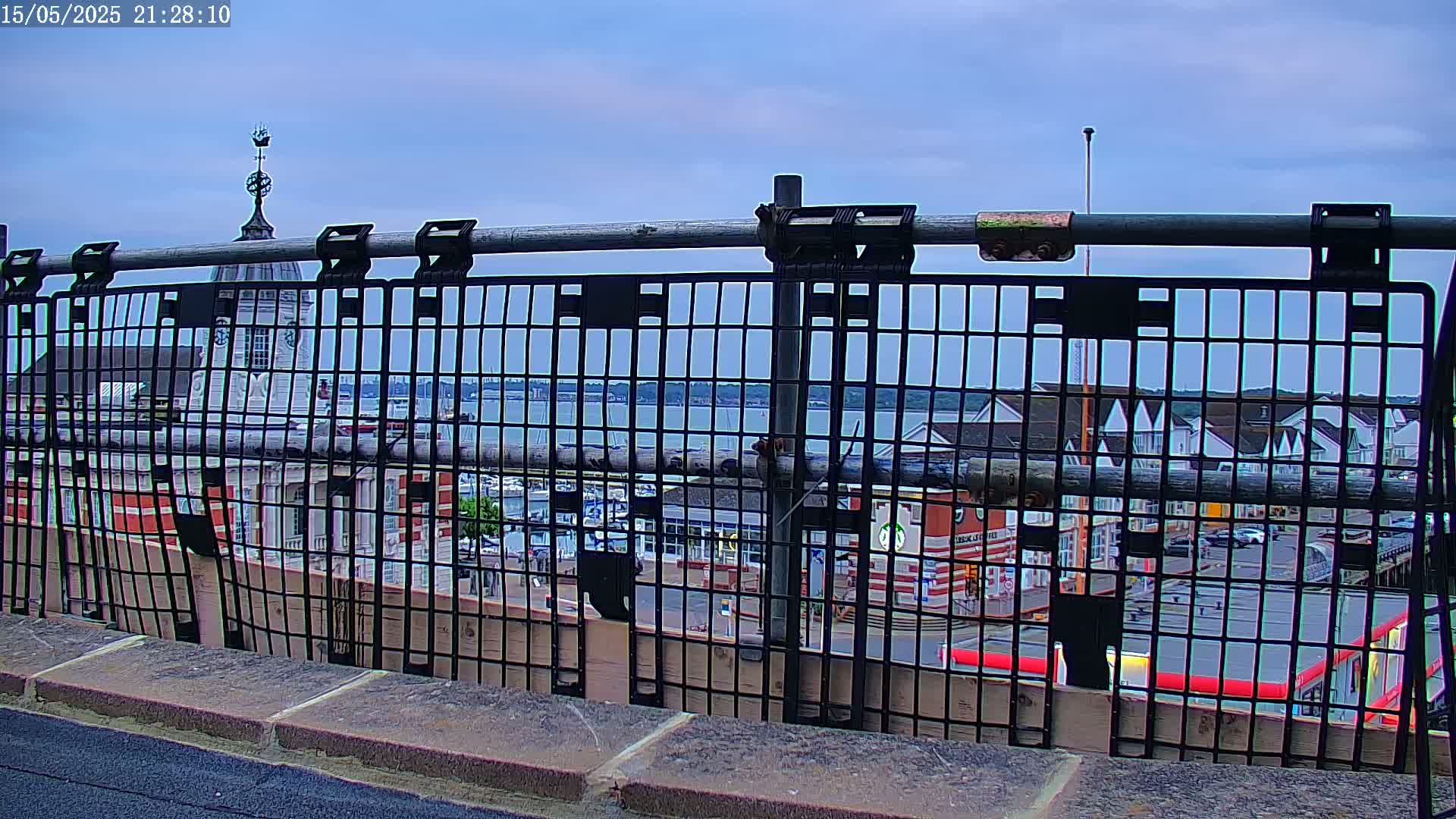 A view through a metal fence shows a waterfront town with buildings, a body of water, and boats under a partly cloudy sky.