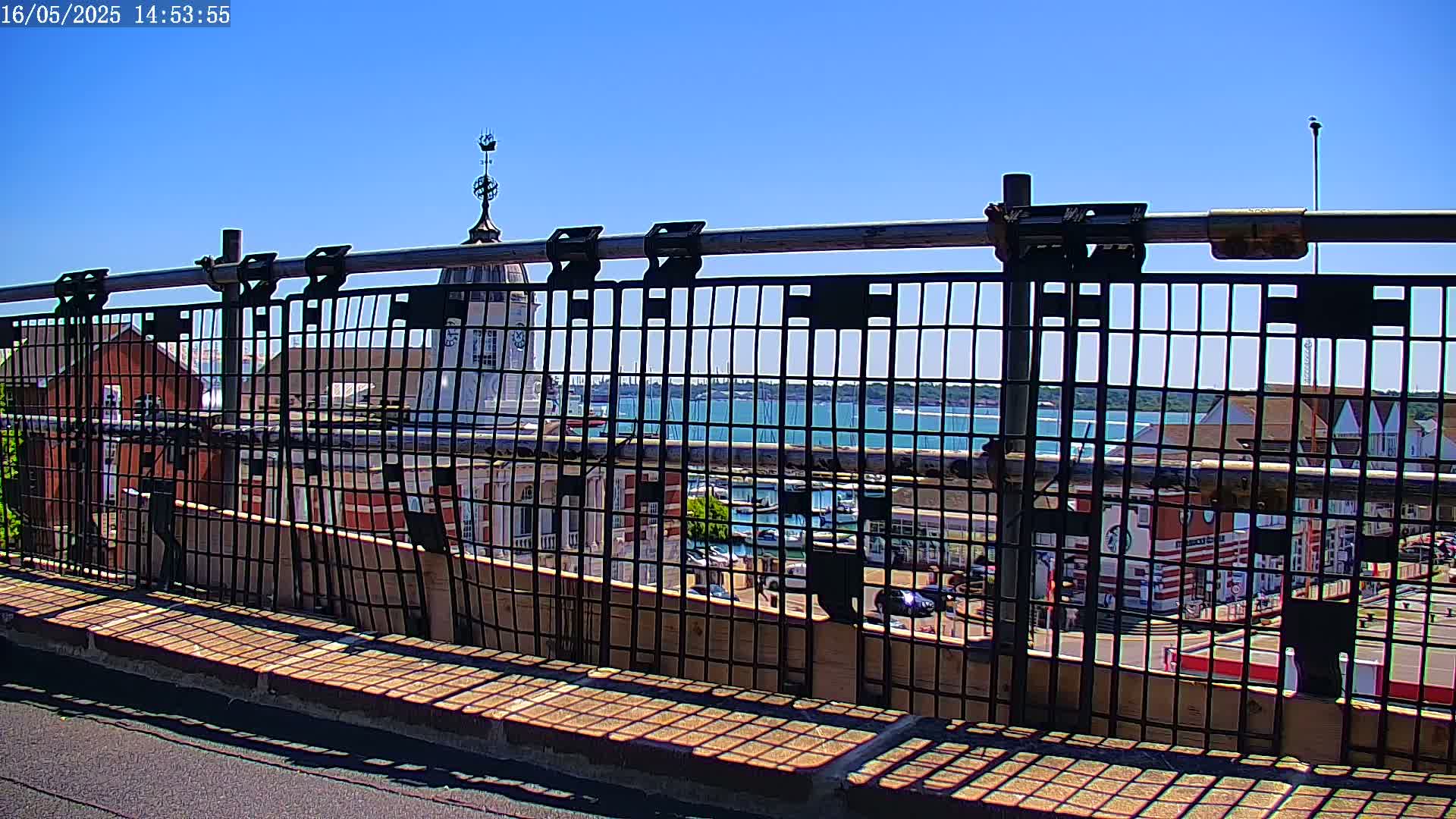 A metal fence provides a view of a harbor with boats and buildings under a clear blue sky.