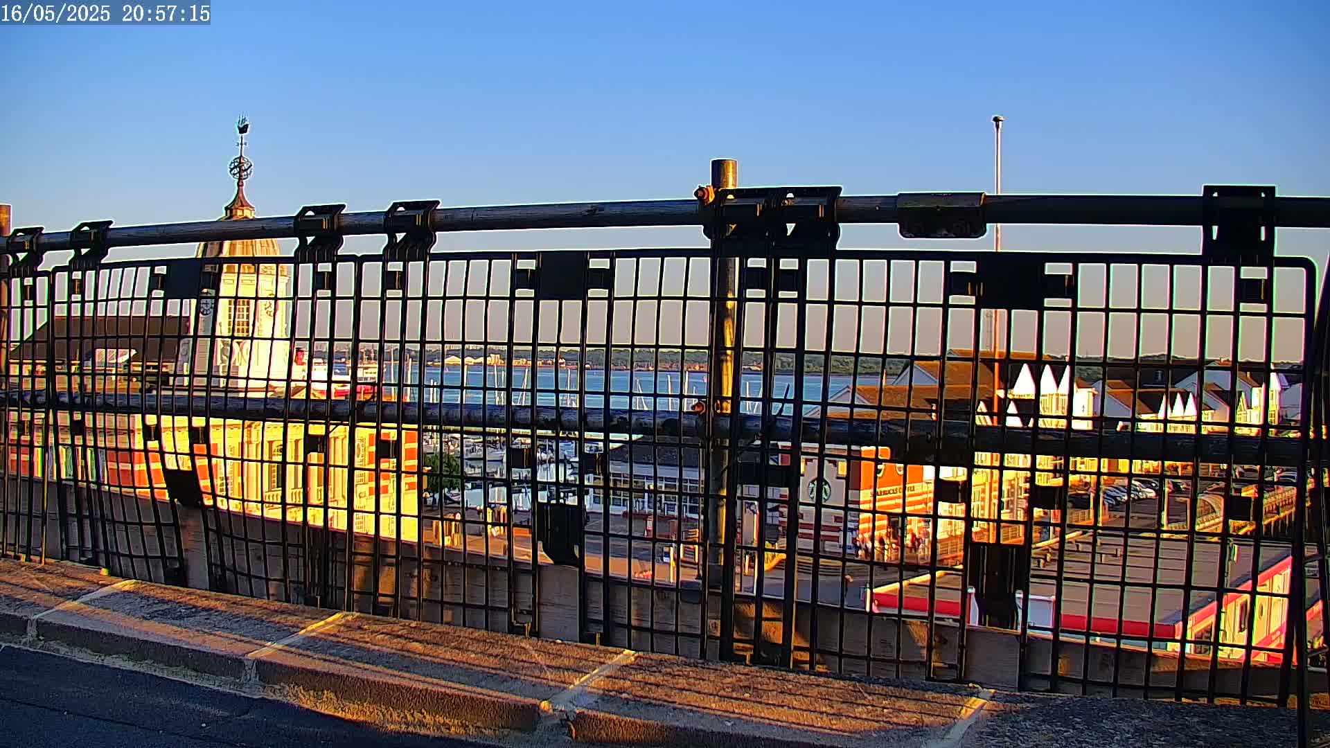 A metal fence obstructs the view of a harbor filled with boats at sunset, with a building featuring a cupola visible in the foreground.