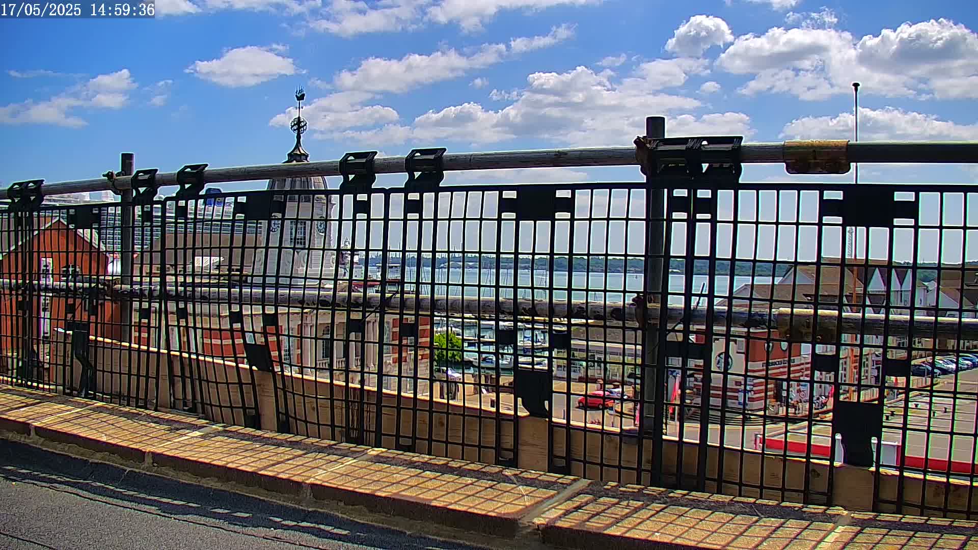 A metal railing provides a view of a harbor with boats, buildings, and a partly cloudy sky.