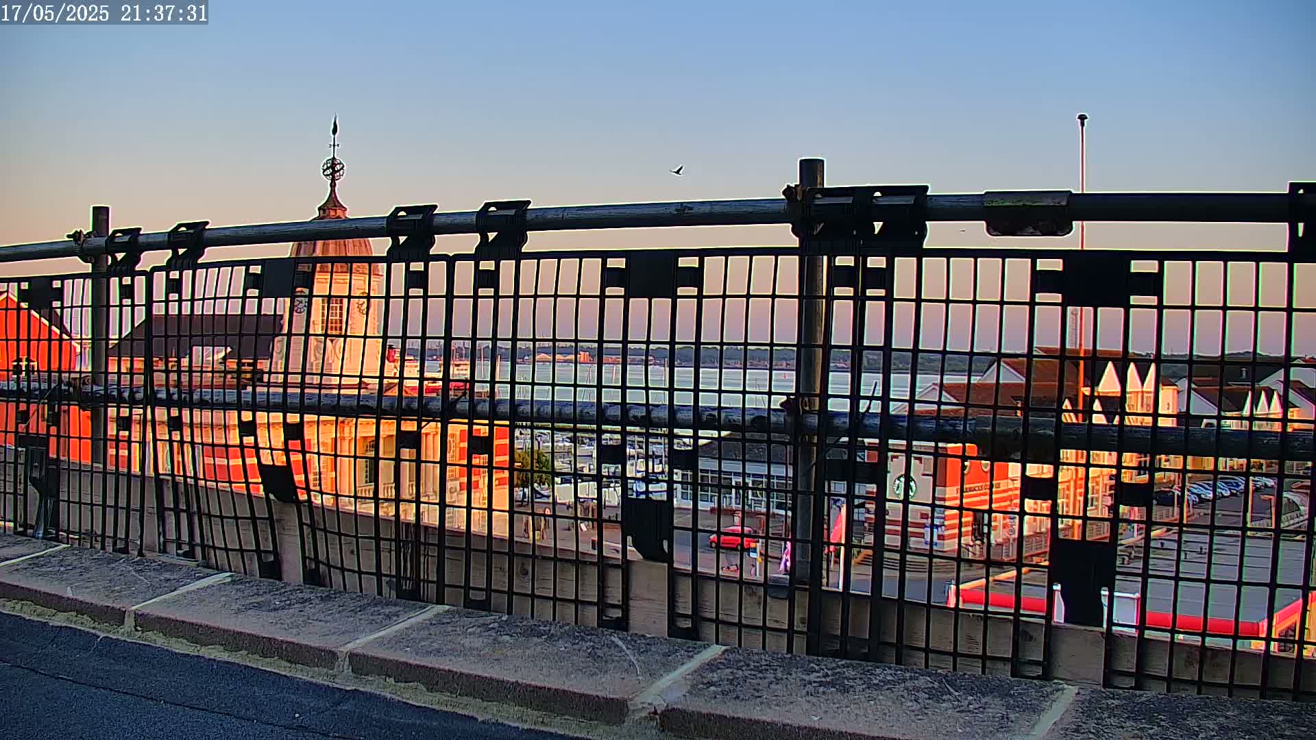 A metal fence partially obscures a view of waterfront buildings at sunset over calm water.