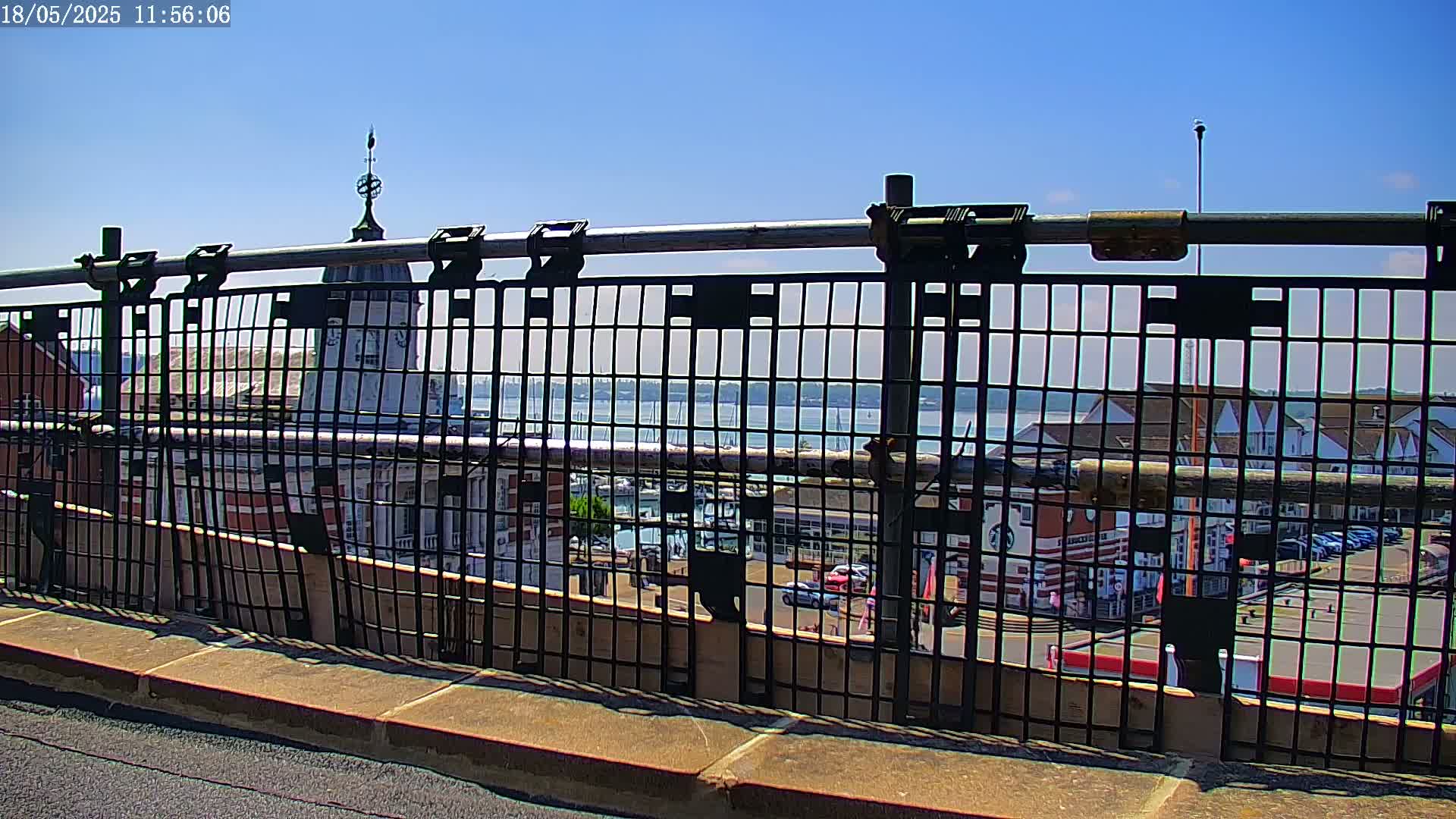 A metal railing overlooks a harbor town on a sunny day, with a body of water and docked boats visible in the background.