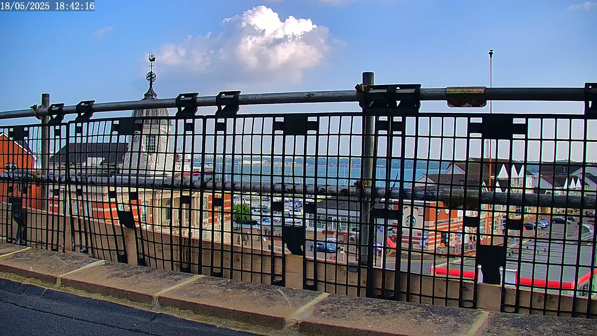 A view of a harbor and town buildings seen through a metal railing on a sunny day with a few clouds.