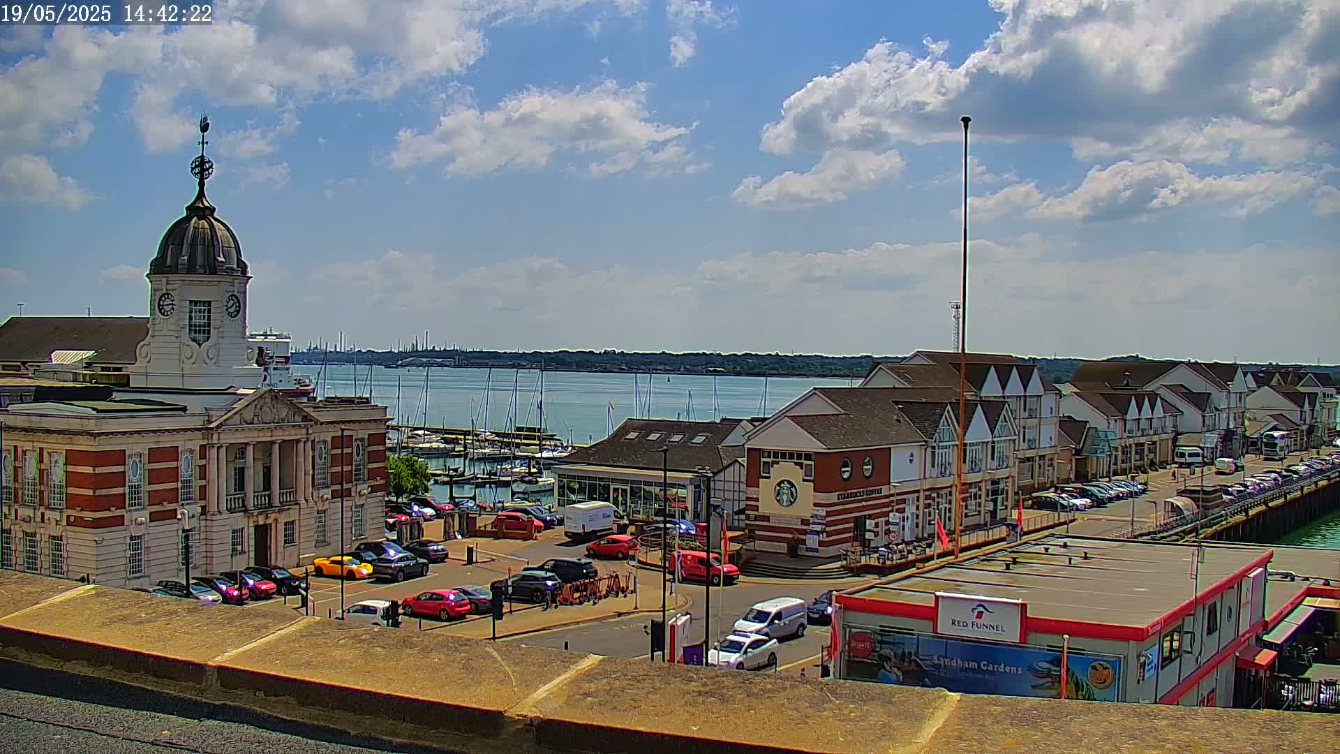 A brick building with a clock tower overlooks a harbor filled with boats under a partly sunny sky.