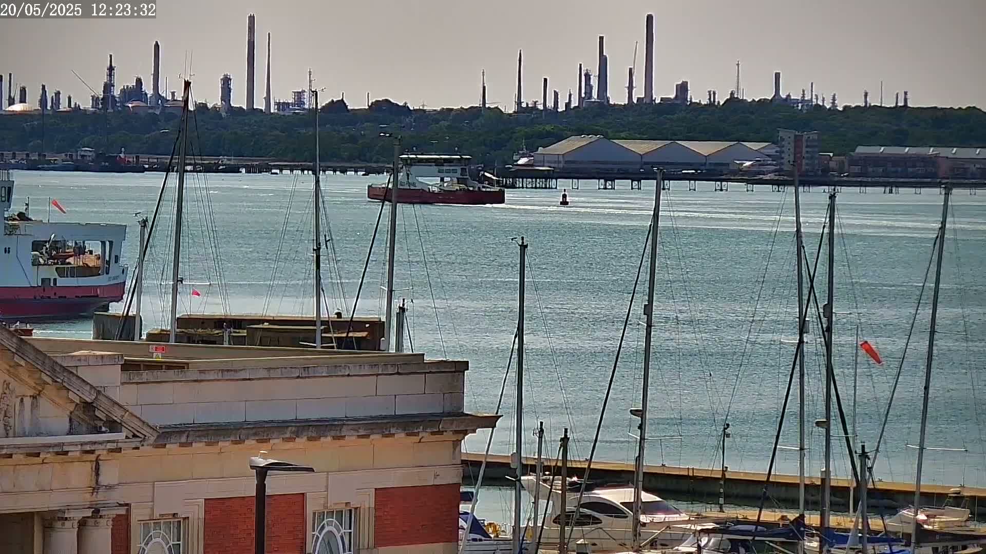 A calm body of water with several sailboats docked near a building, a ferry, and a cargo ship; in the background, an industrial refinery is visible under a hazy sky.