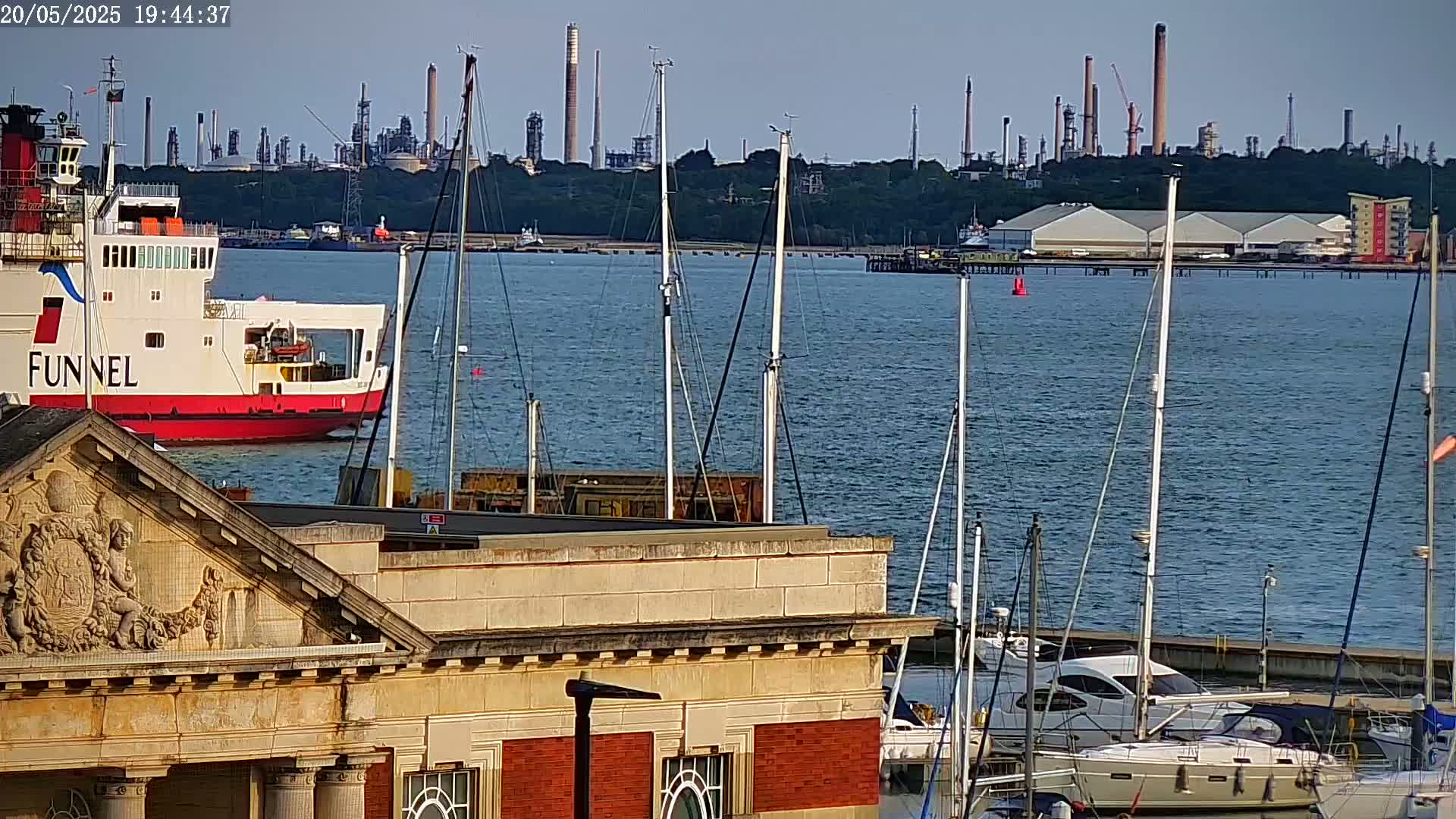 A ferry is docked in a harbor with numerous sailboats moored nearby, an industrial plant is visible in the background across the calm water.