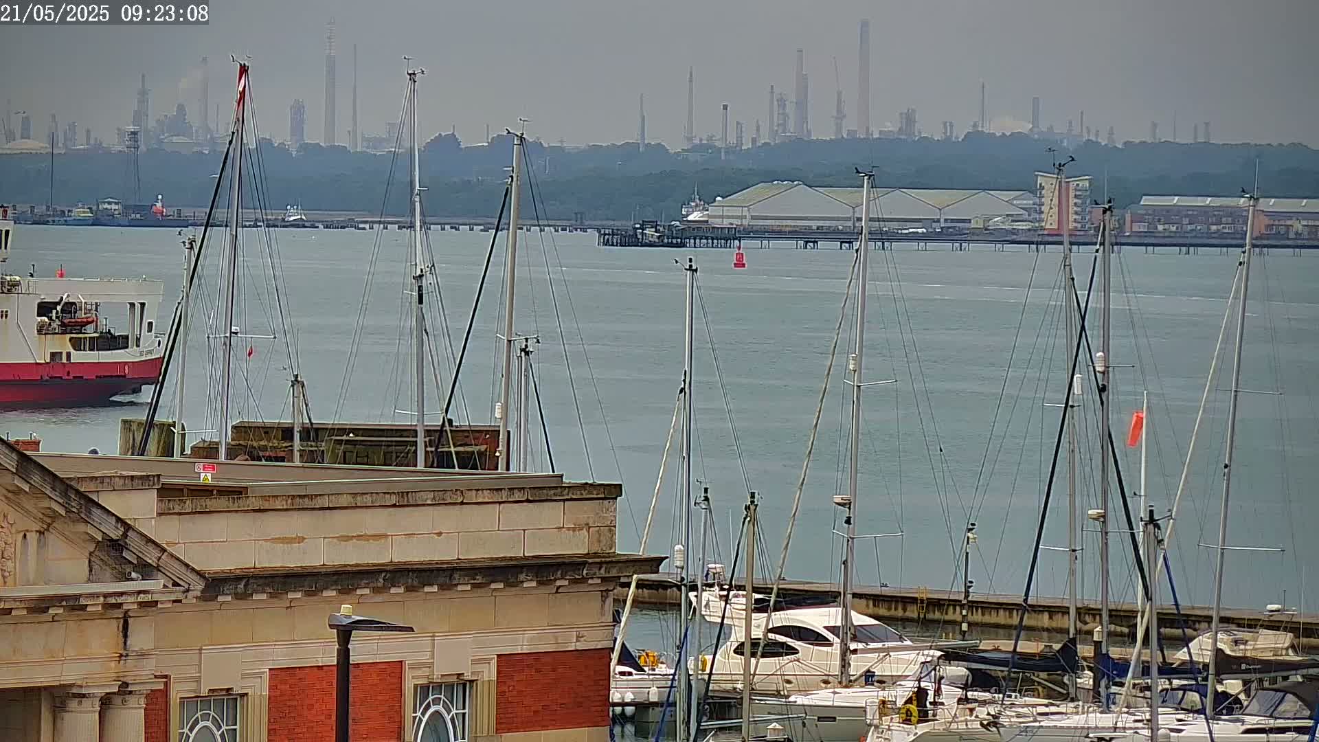 A calm body of water shows a marina with numerous sailboats docked, a ferry in the distance, and an industrial area visible in the background under an overcast sky.