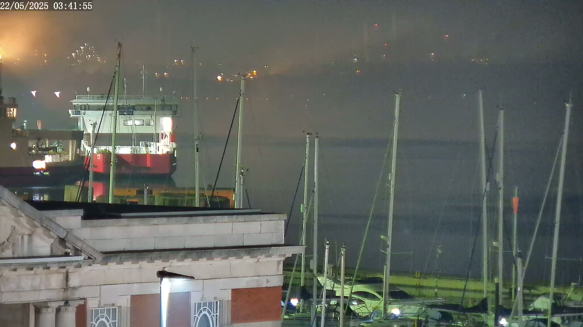 A red barge is moored in a harbor at night in foggy conditions, with numerous sailboats masts visible in the foreground and faint lights in the background.