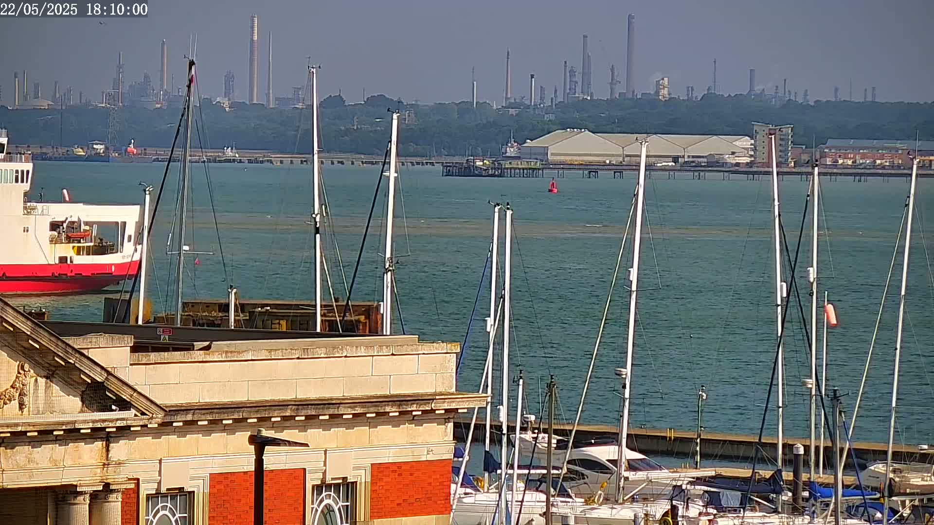 A red ferry is moored near a marina filled with sailboats, with an industrial complex visible in the background on a sunny day.