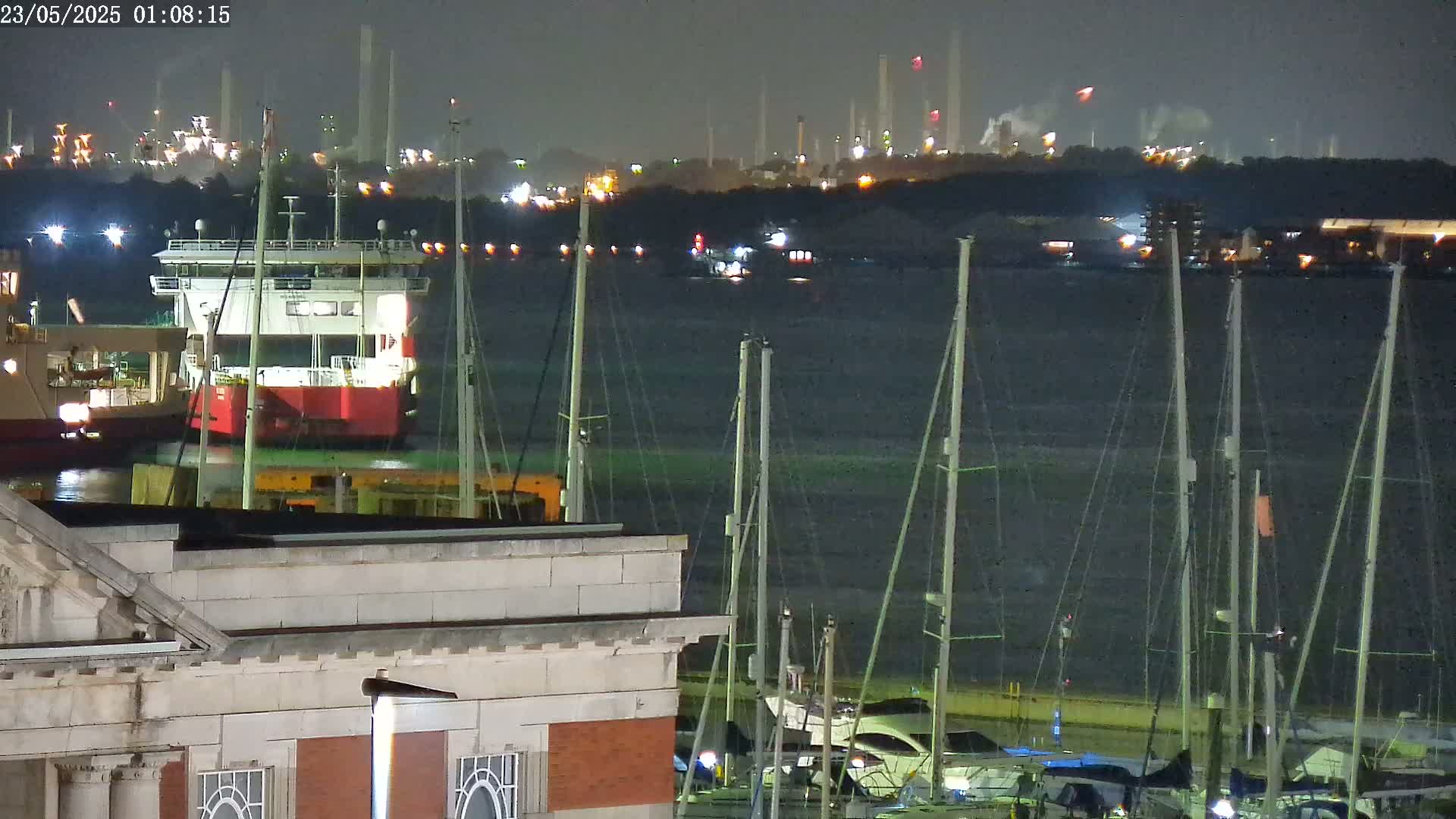 A nighttime view of a harbor with docked sailboats in the foreground, a red barge in the midground, and an industrial area with lights and smoke stacks in the background.