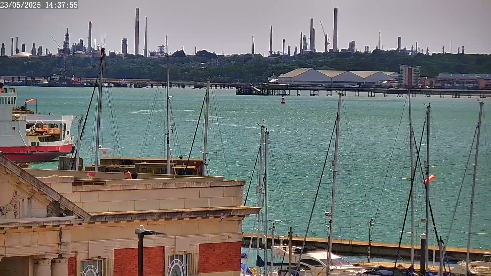 A ferry and sailboats are docked near a building overlooking a body of water with a large industrial plant visible in the distance on a sunny day.