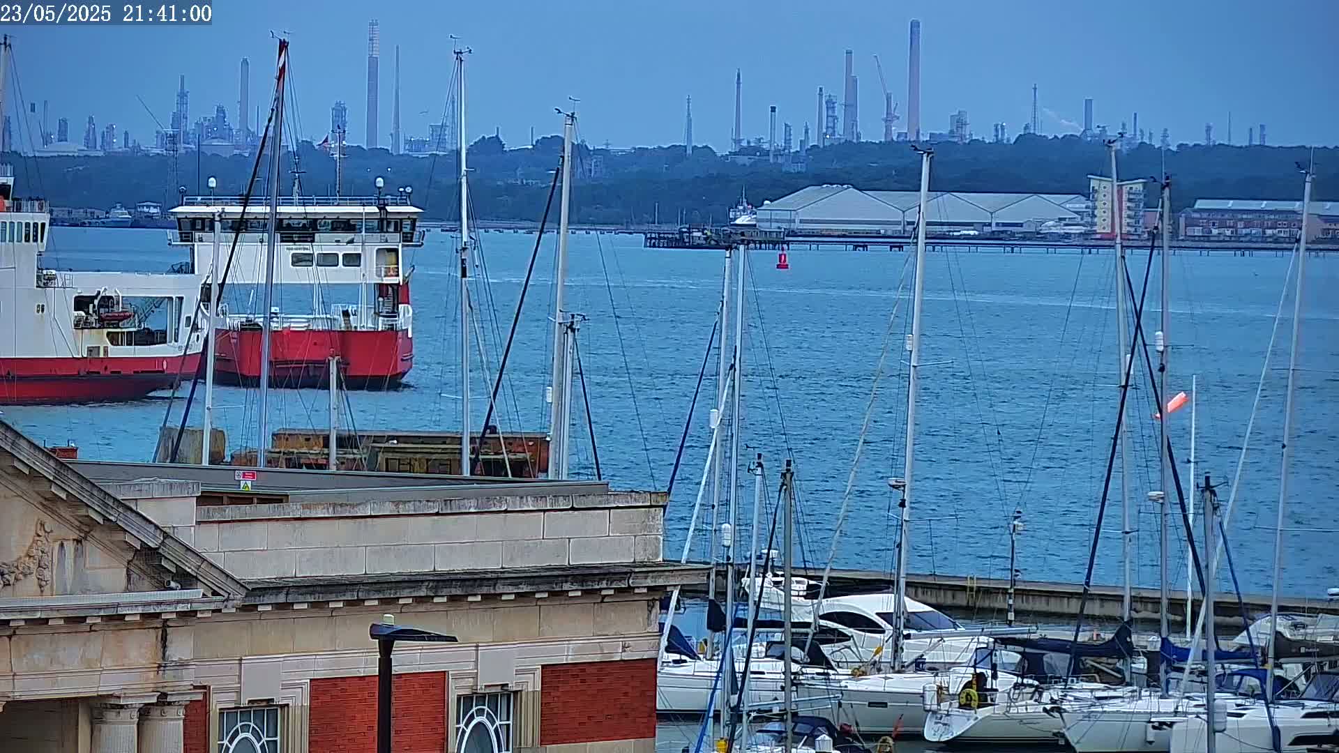 A calm body of water shows a ferry and numerous sailboats docked at a marina, with an industrial skyline visible in the distance.