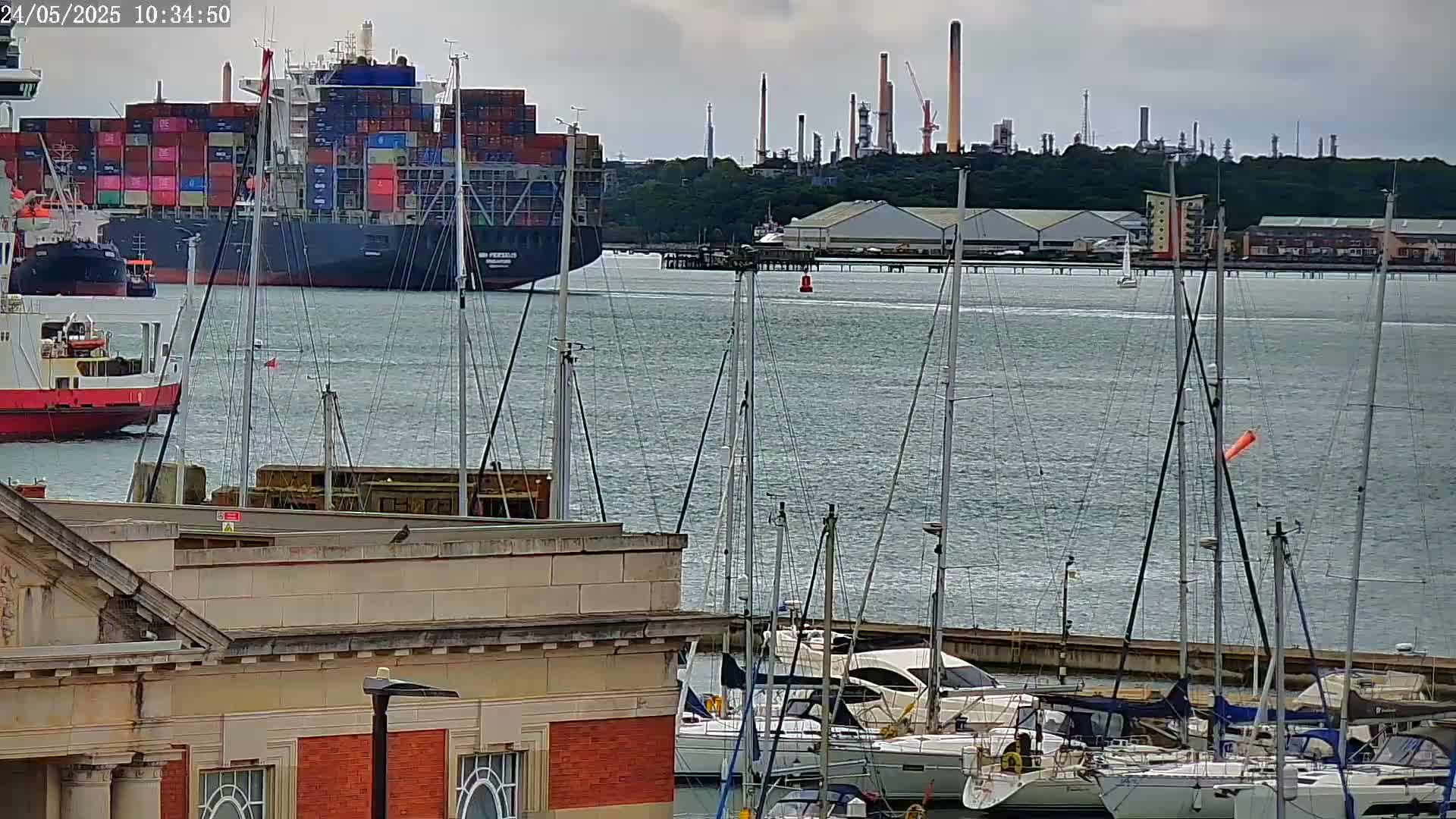A large container ship and several sailboats are in a harbor on an overcast day, with an industrial area visible in the background.