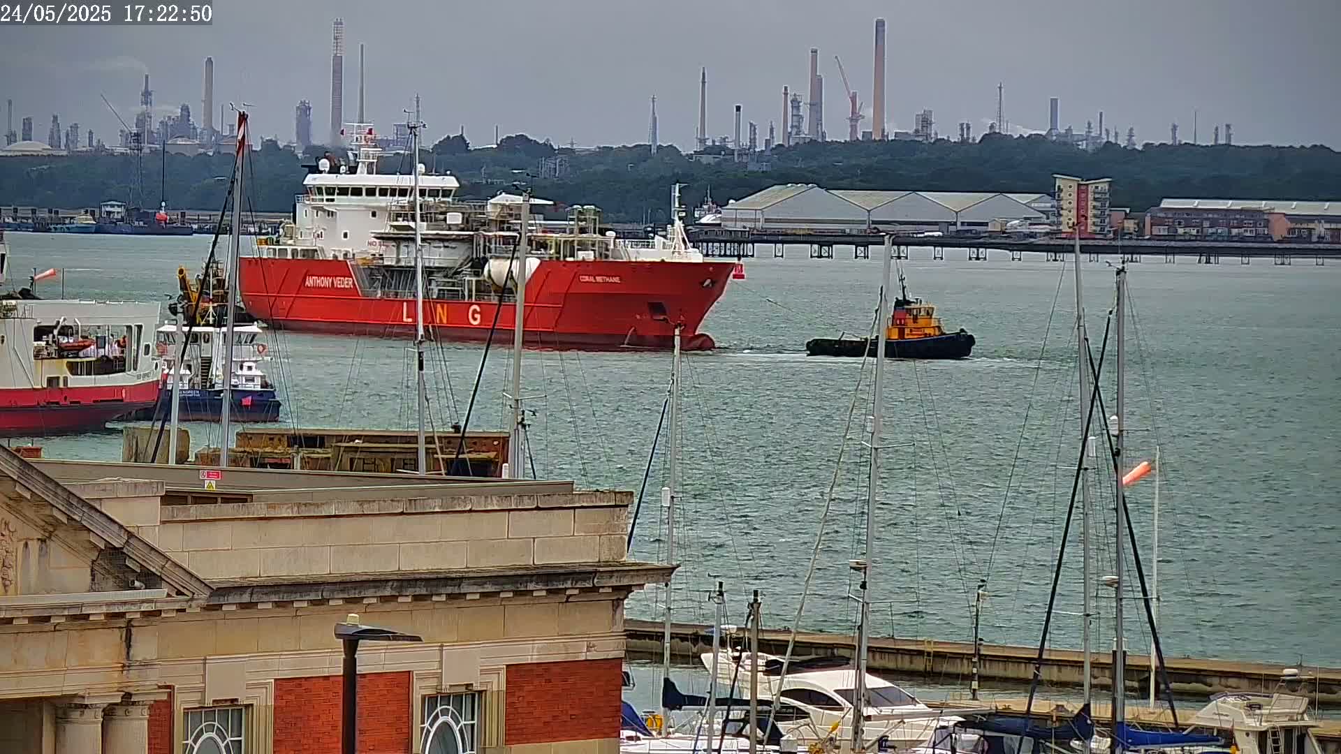 A large red ship is being towed by a smaller tugboat in a calm body of water, with several docked sailboats and other ships in the foreground and an industrial refinery in the background.