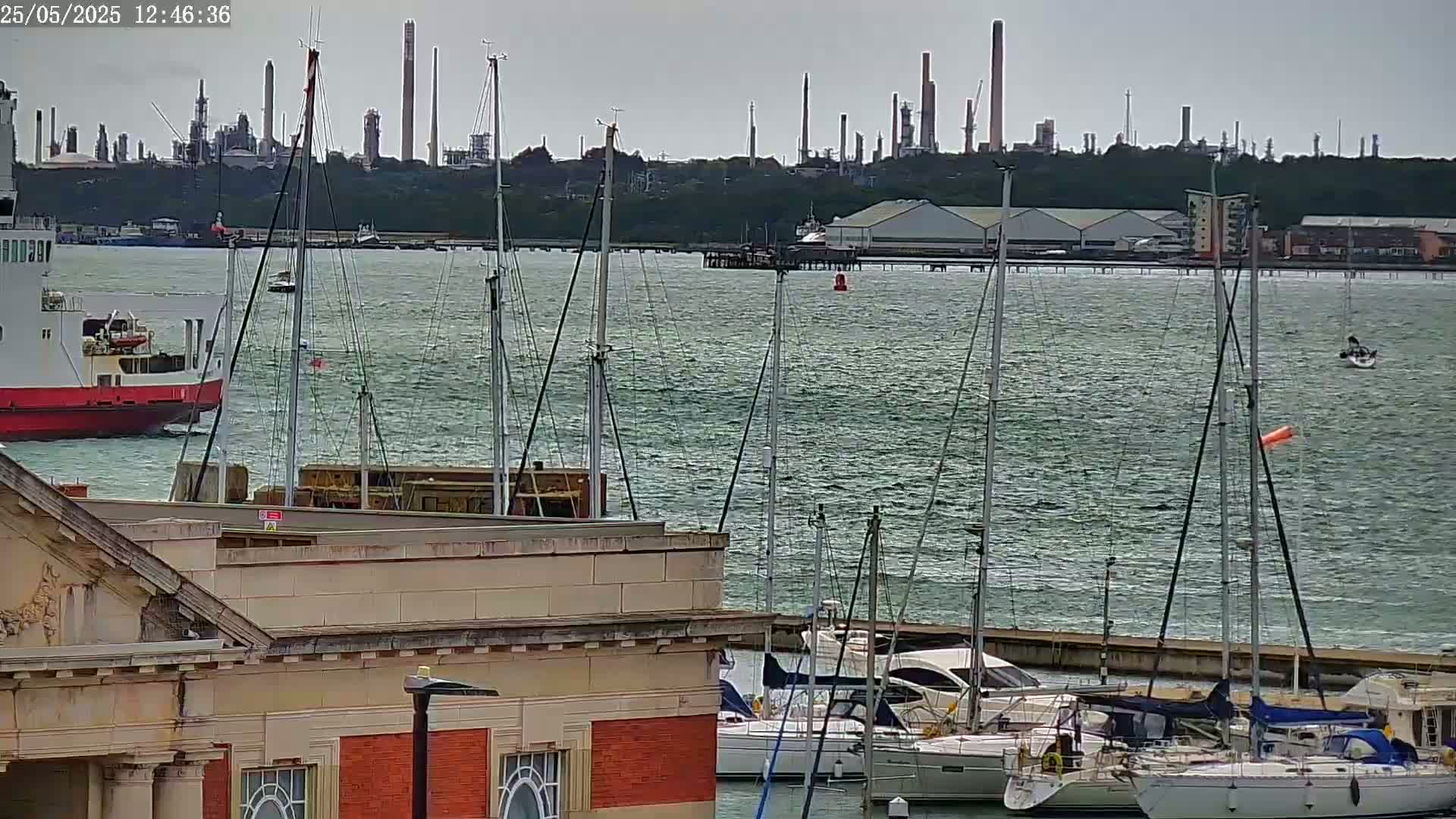 A red ferry is docked near a marina filled with sailboats on a choppy, gray day with an industrial plant visible in the distance.