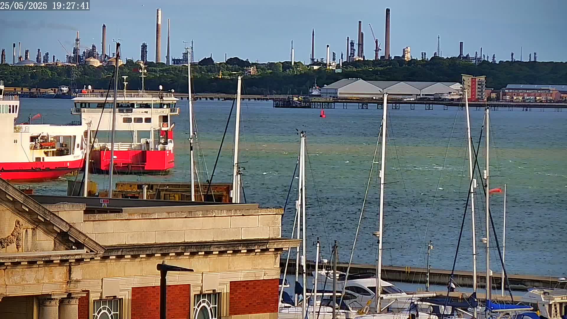 Two red ferries are docked in a harbor next to a building, with sailboats moored in the foreground and an industrial refinery in the distance on a sunny day with calm water.