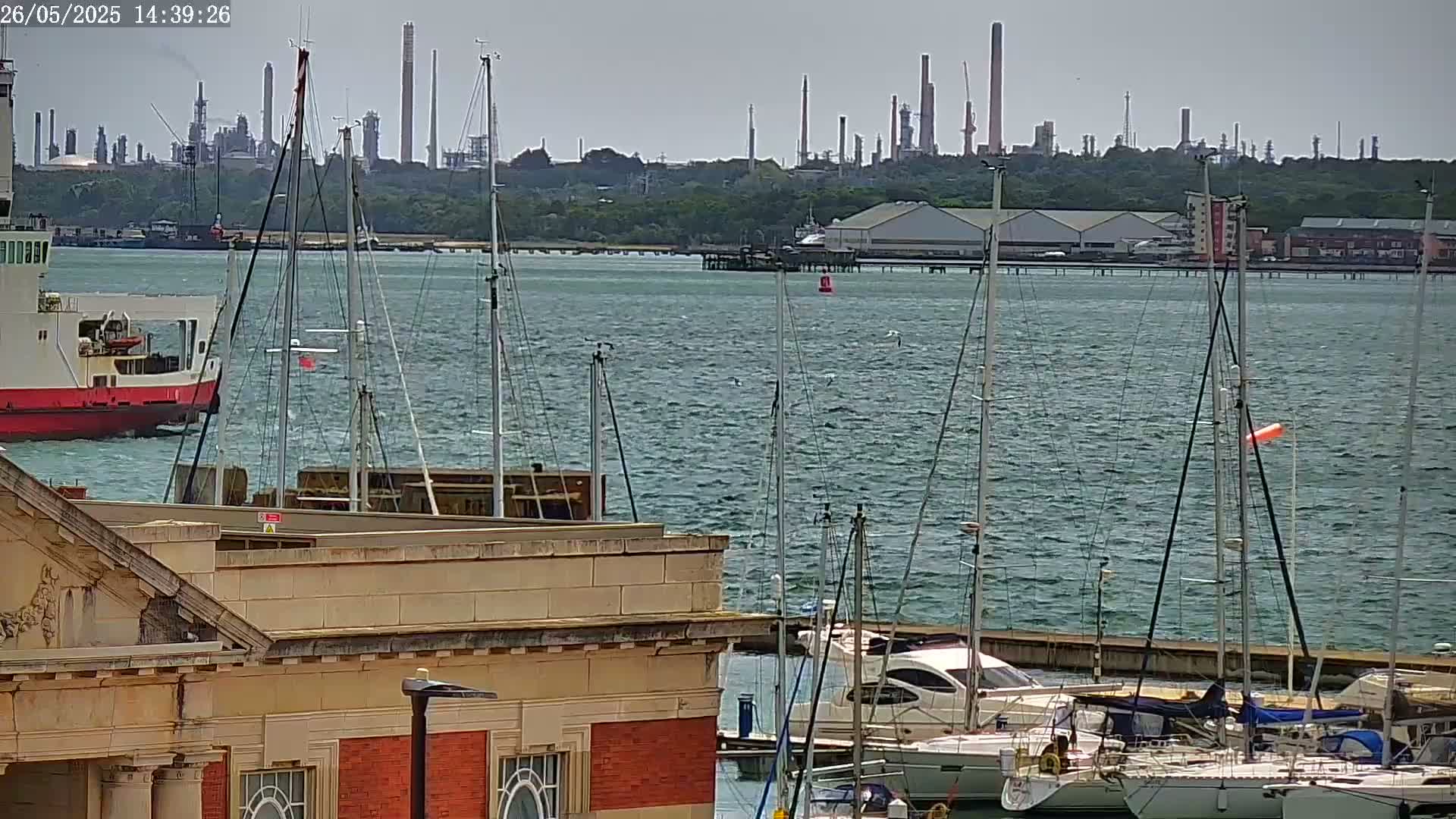 A view of a harbor with sailboats and a ferry, a distant industrial complex, and a building in the foreground, under somewhat cloudy skies.