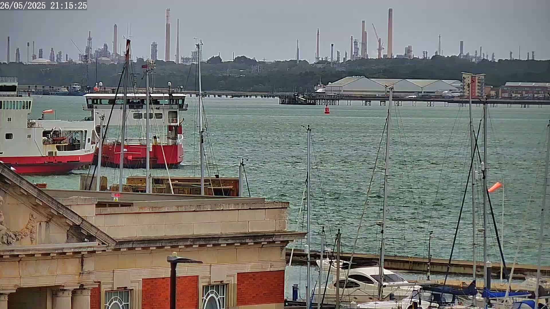 A harbor scene with two ferries, numerous sailboats moored at a dock, and an industrial plant visible in the distance on an overcast day.
