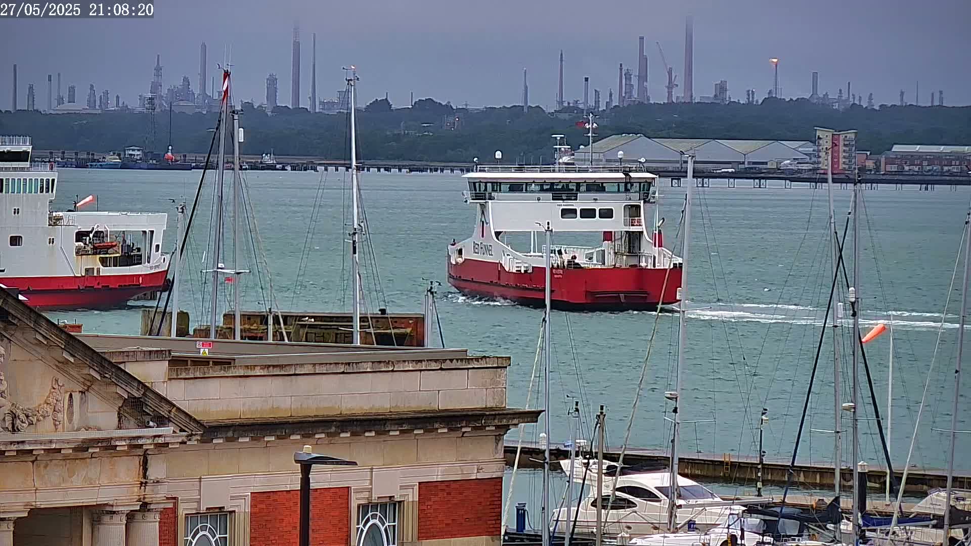 A red ferry sails past a marina on an overcast day, with an industrial plant visible in the distance.
