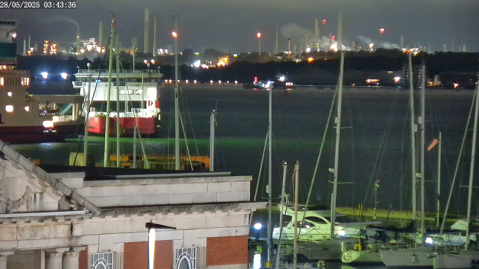 At night, a harbor with docked sailboats is seen in front of a large cargo ship and an industrial plant with illuminated structures and smoke.