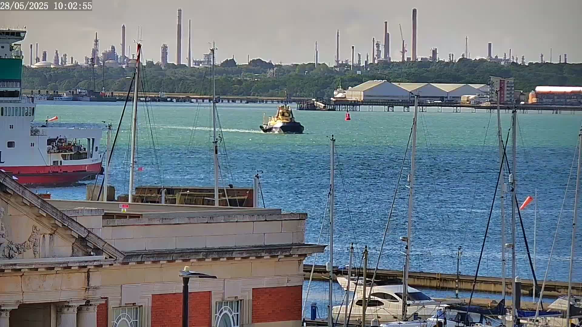 A tugboat moves across a calm, blue body of water, past a docked ferry, with a refinery and marina visible in the background.