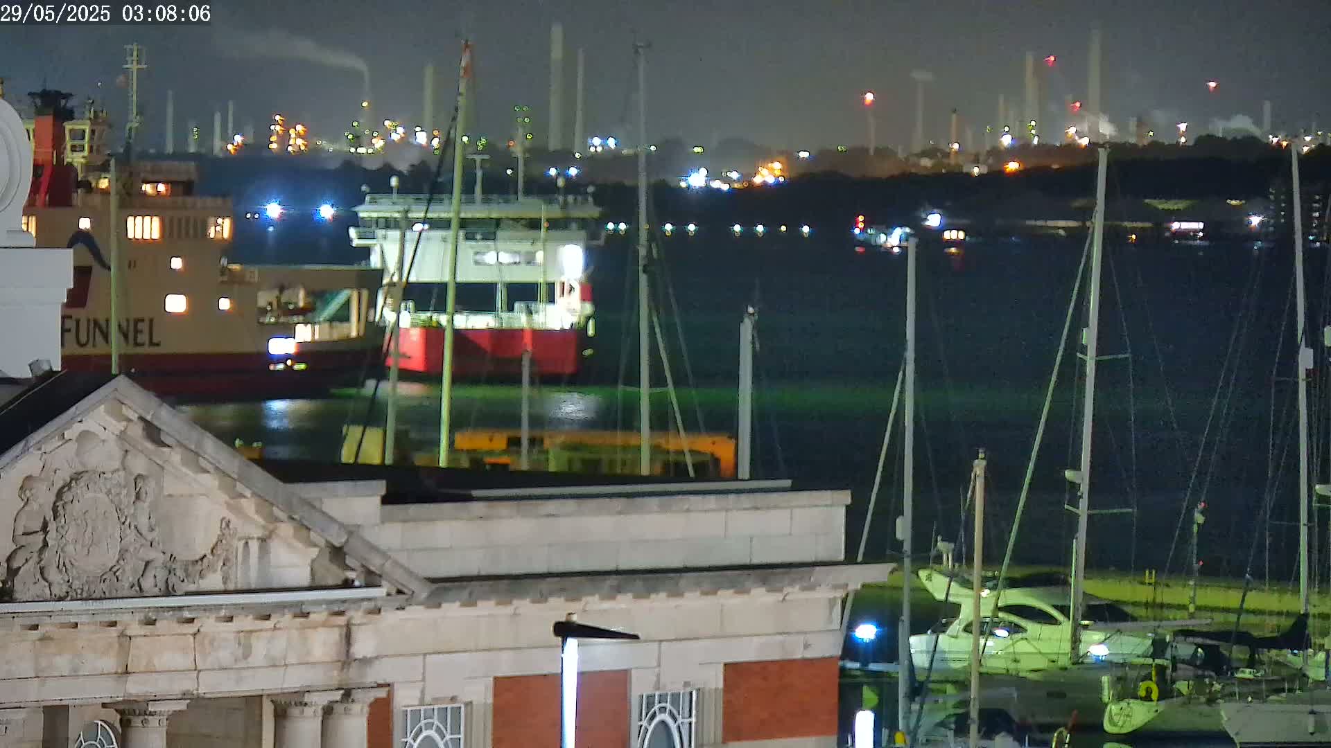 At night, a large cargo ship and several sailboats are moored near a stone building with ornate detailing, with an industrial plant illuminated in the background.