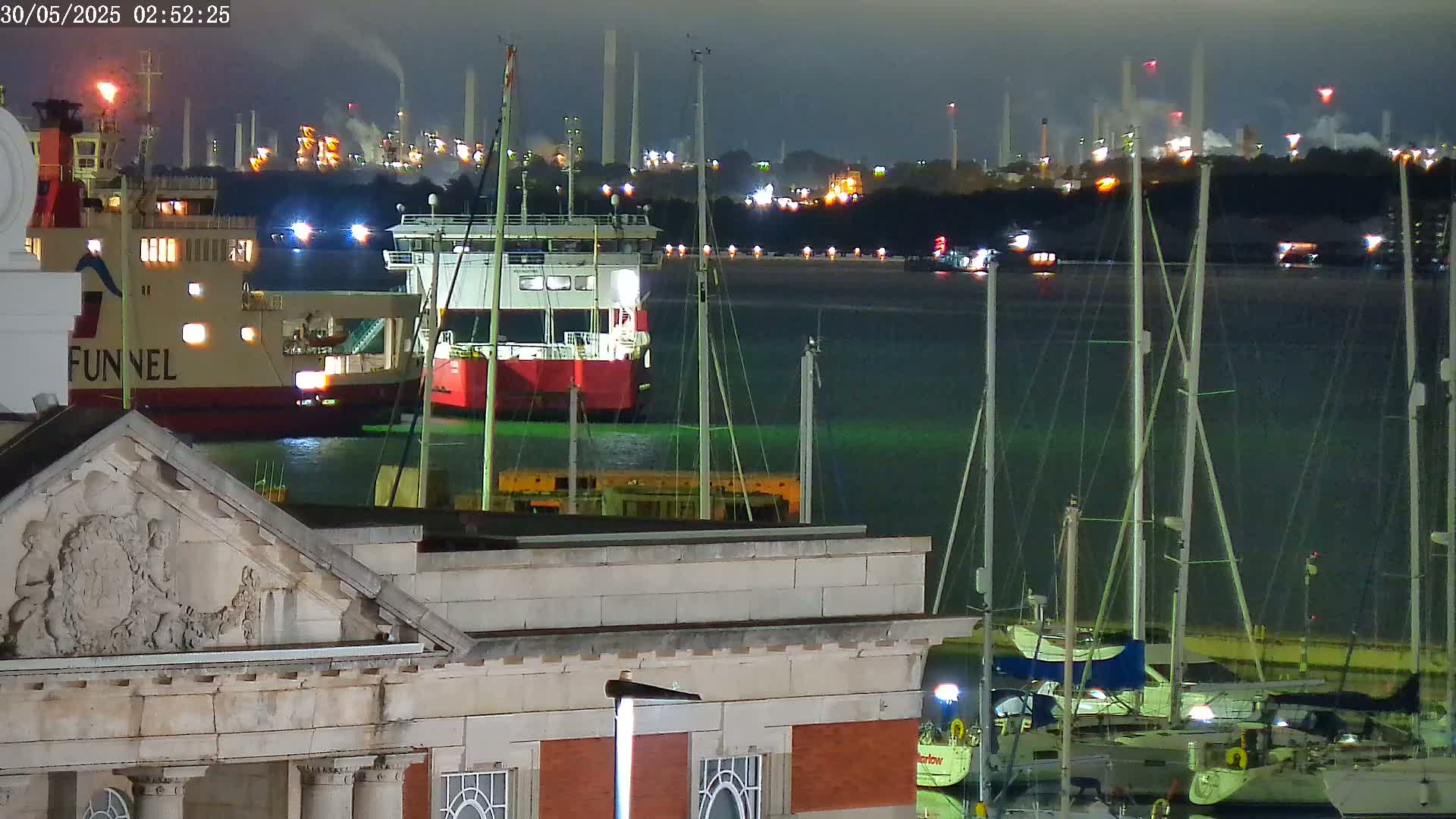 At night, a large ferry and several sailboats are moored in a harbor, with an industrial plant glowing in the distance.