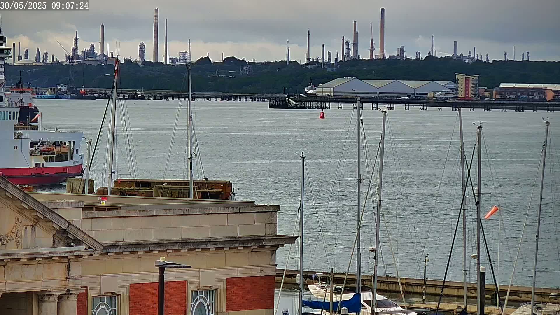A calm, overcast day shows a body of water with docked sailboats in the foreground, a large cargo ship, and an industrial refinery in the background.