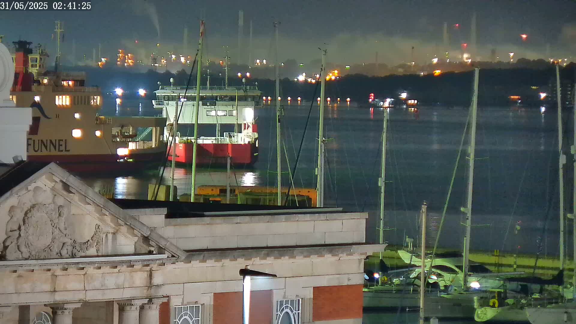 At night, a ferry and several sailboats are moored in a harbor, with an industrial area faintly visible in the background under a clear sky.