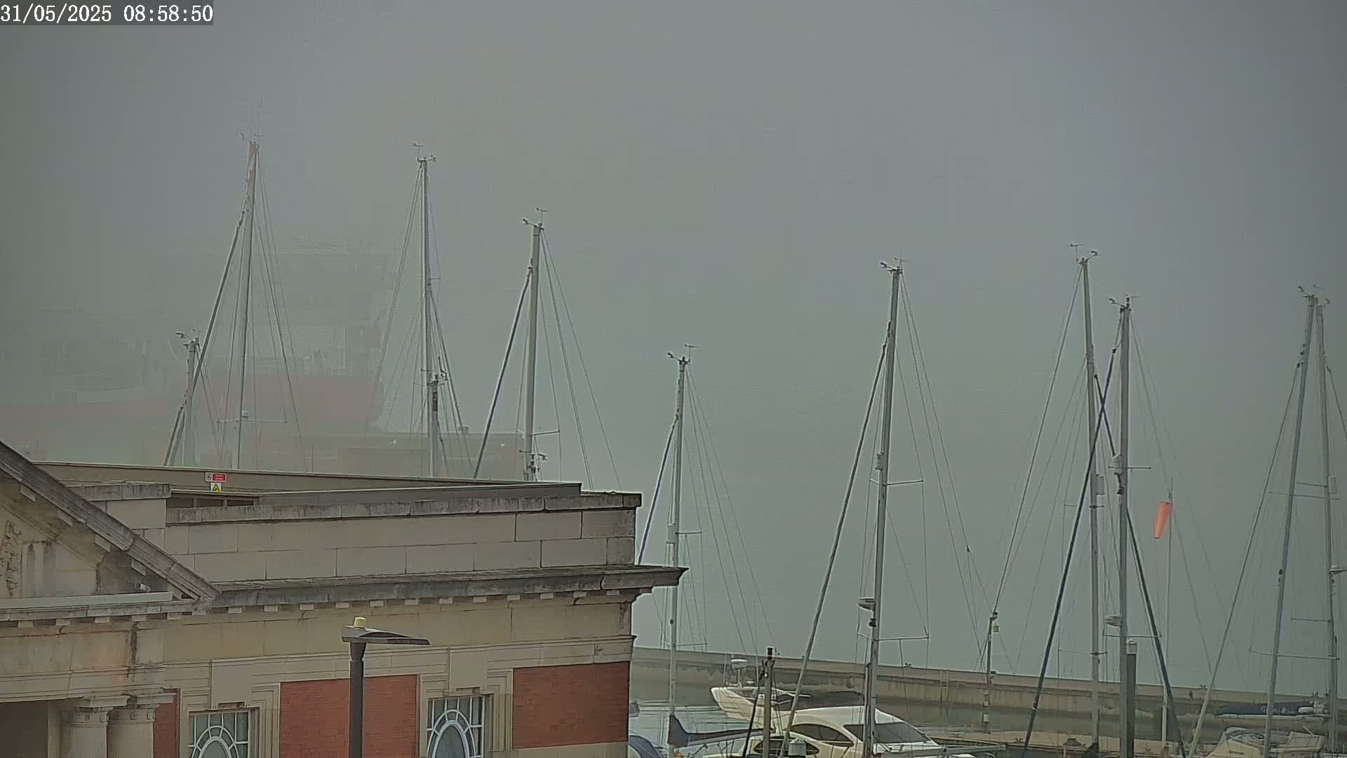 A foggy harbor scene shows several sailboat masts rising above a low stone building.
