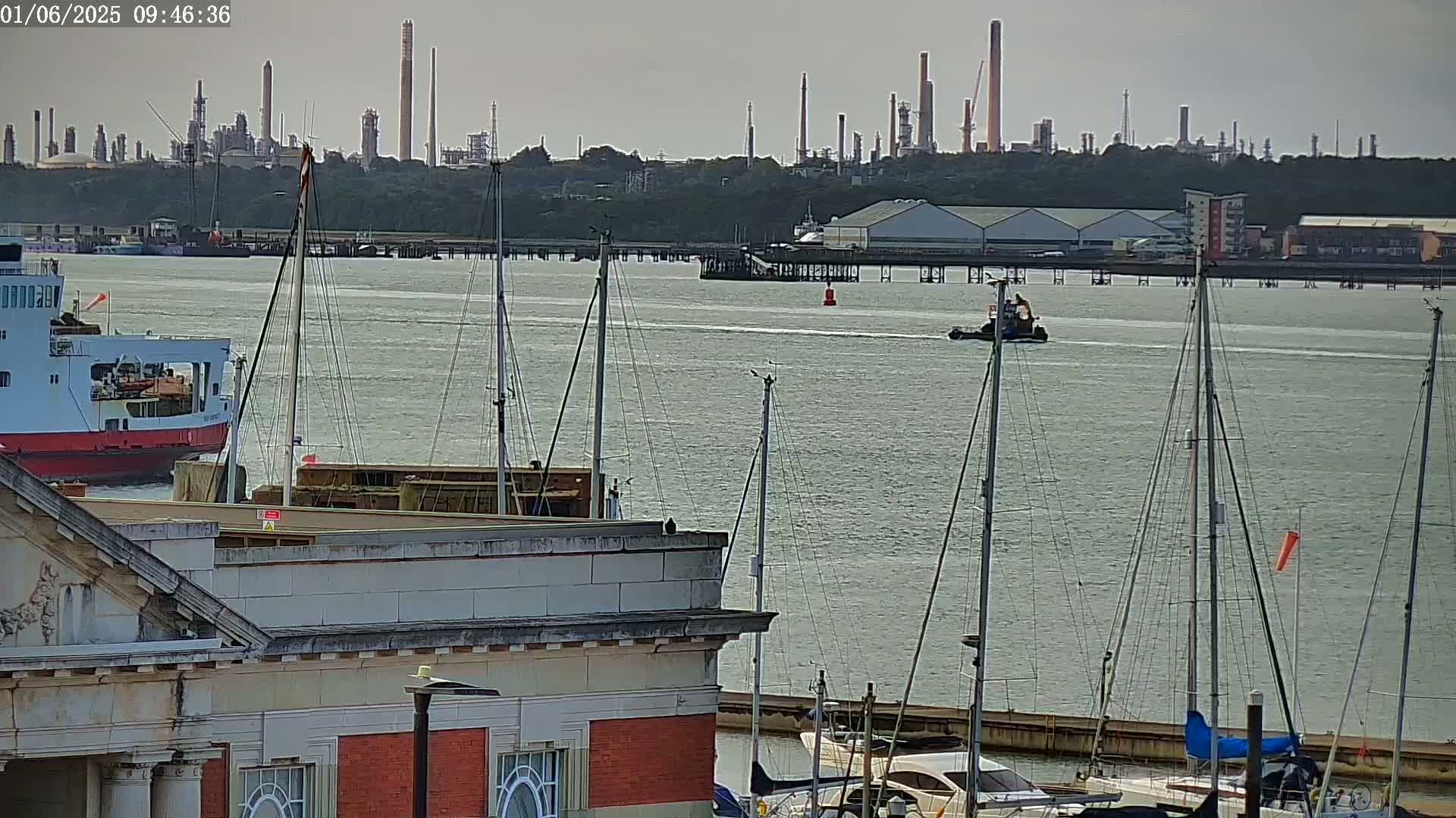 A tugboat and a ferry are on a calm body of water with numerous sailboats docked in the foreground, and an industrial refinery in the background.