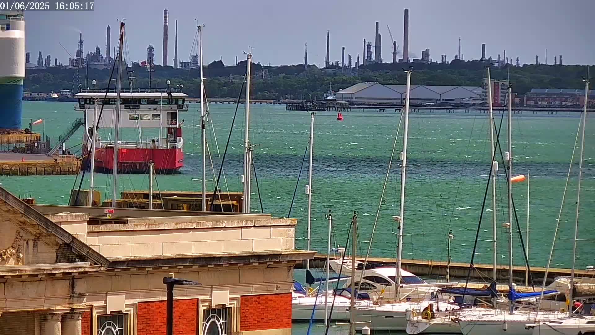 A red ferry is docked near a marina filled with sailboats on a sunny day with a slightly choppy teal-green sea, and an industrial plant is visible in the background.