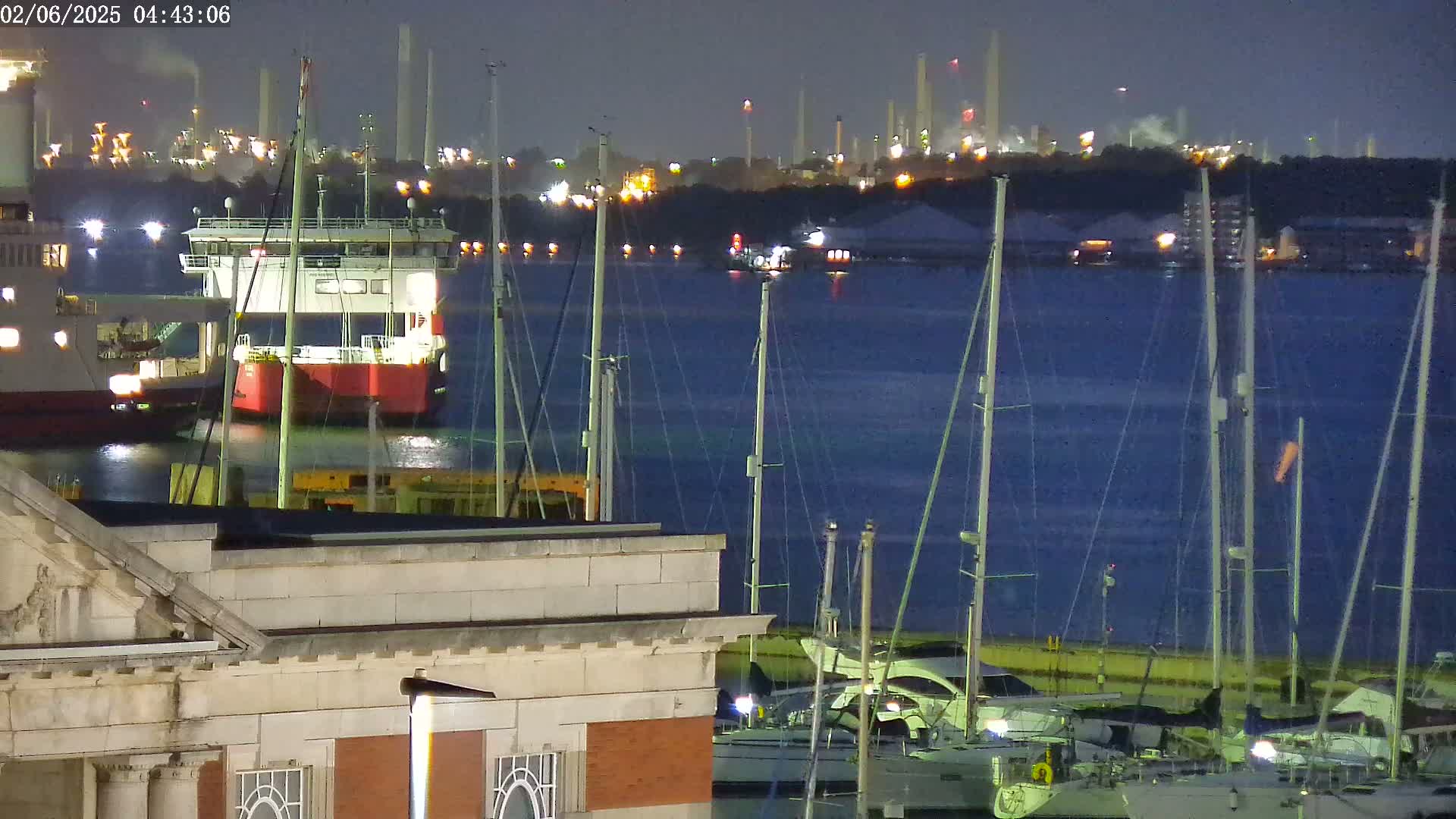 A nighttime harbor scene shows a docked barge and several sailboats, with an industrial complex faintly illuminated in the background under a dark sky.