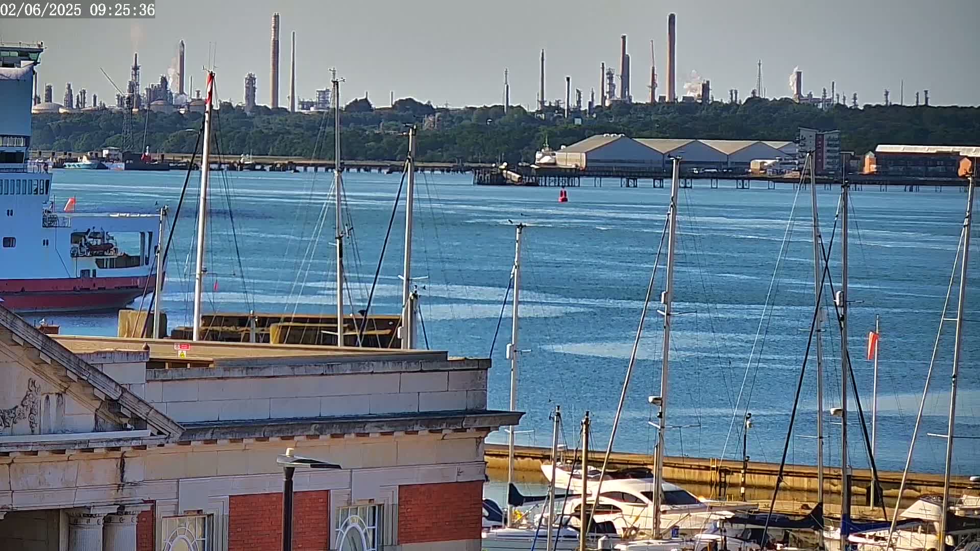 A ferry is docked near a marina filled with sailboats, with an industrial refinery visible in the distance on a sunny day.