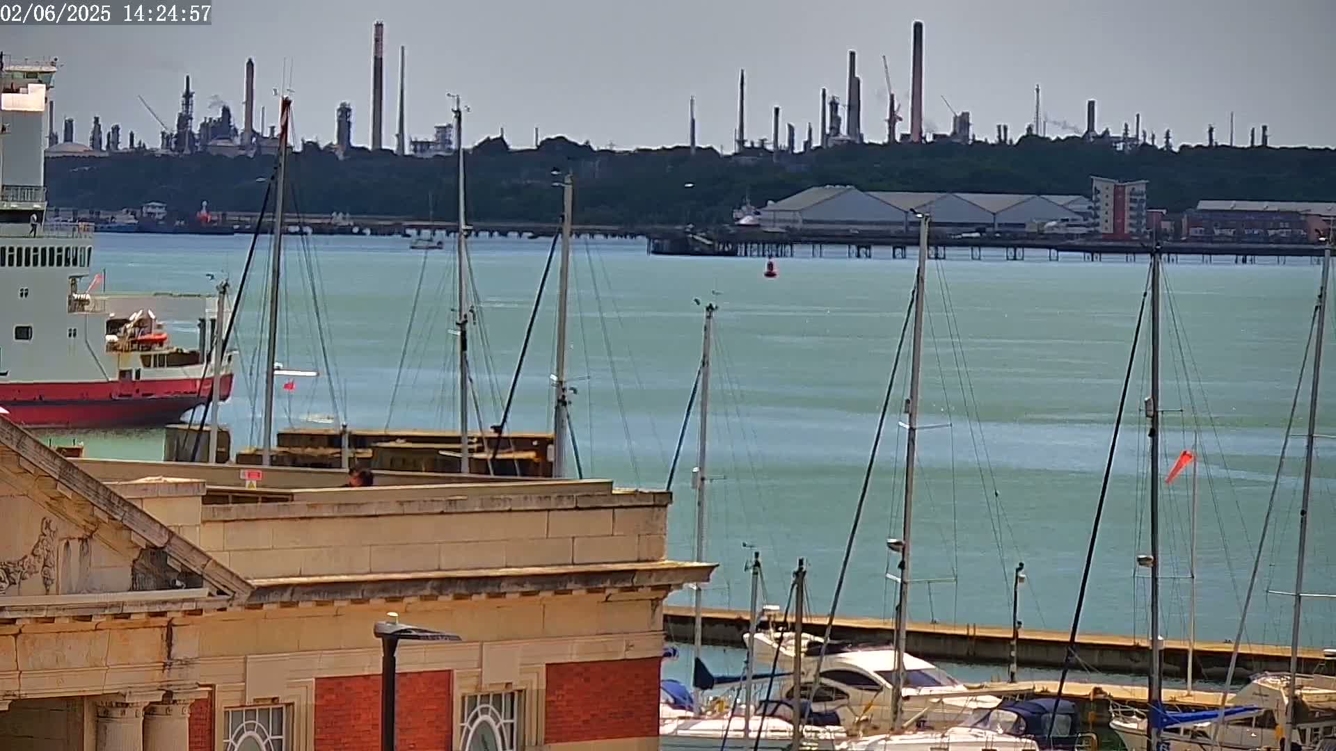A red ferry is docked near a marina full of sailboats, with an industrial plant visible in the distance across a calm, teal-colored body of water under a hazy sky.