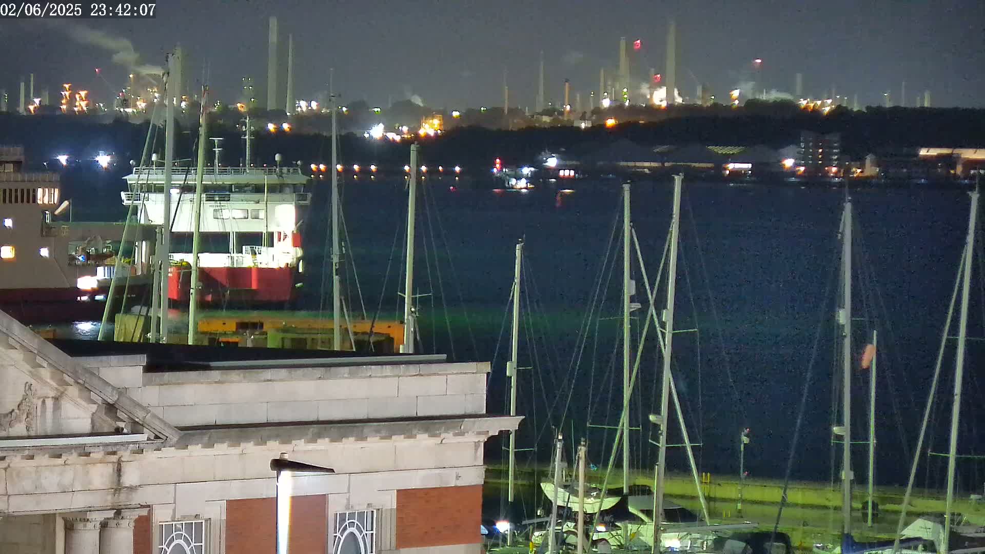 A nighttime view of a harbor with docked sailboats in the foreground, a barge in the middle ground, and an industrial plant illuminated on the horizon under a dark sky.