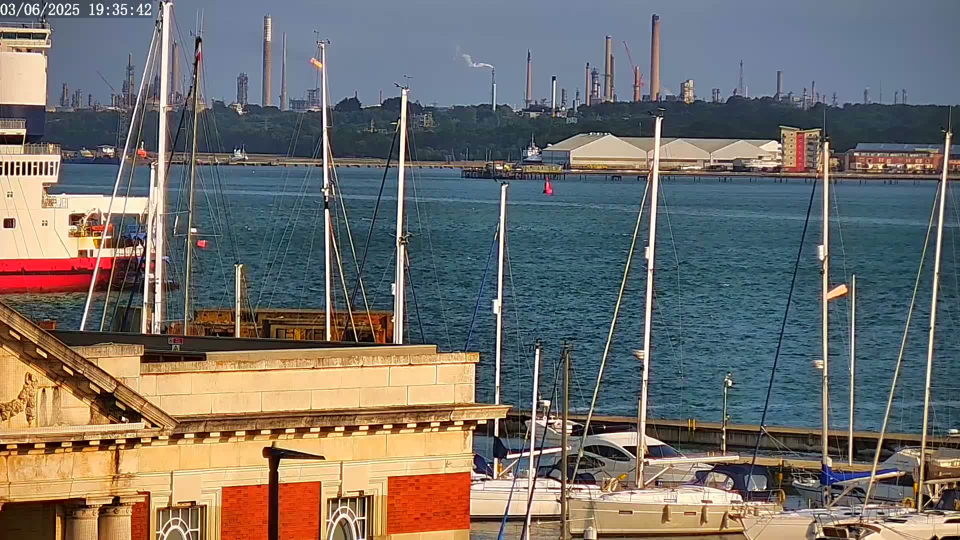 A calm body of water with numerous sailboats docked near a brick building, featuring a large ship and an industrial refinery in the background under a clear sky.