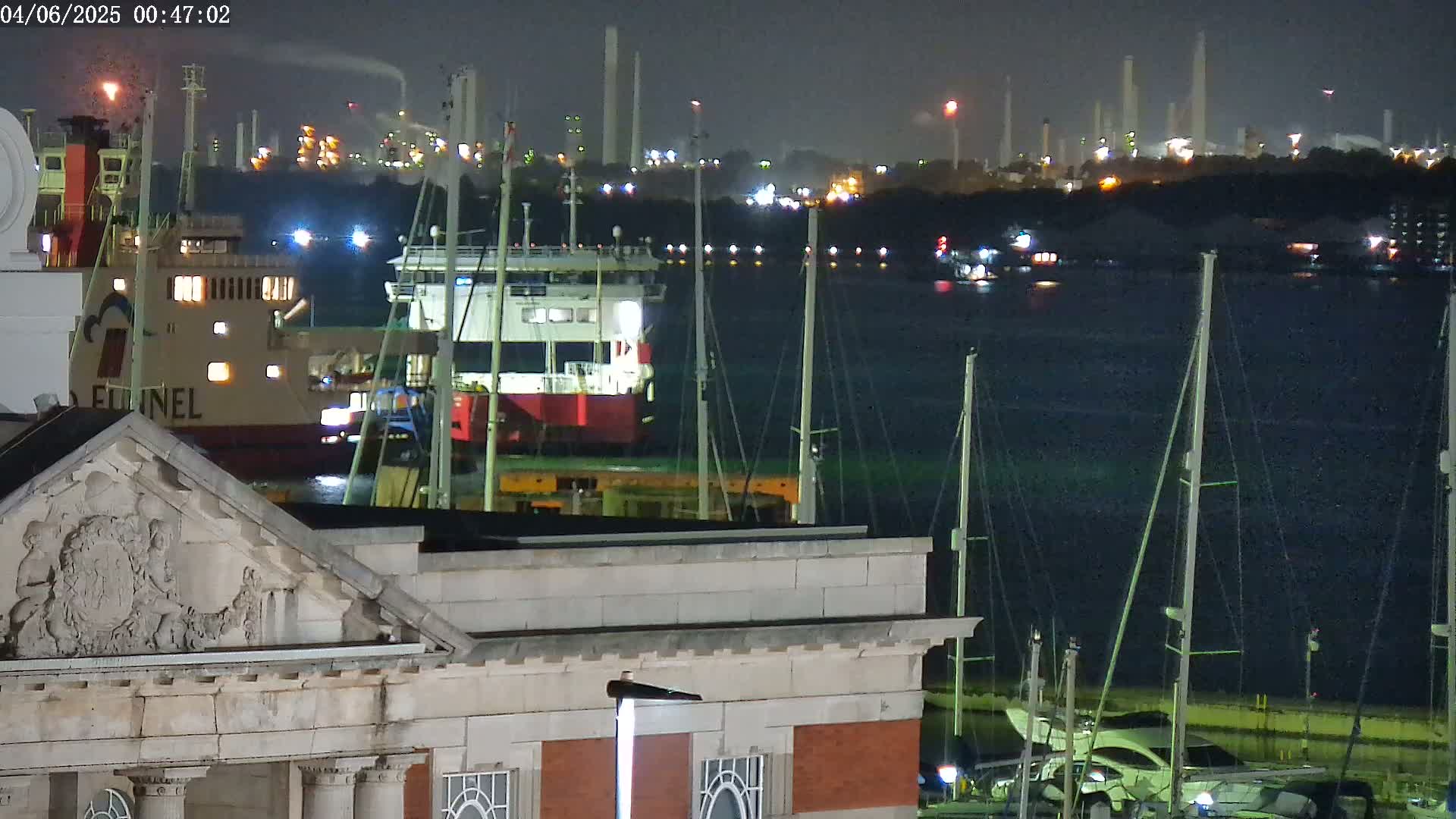 At night, a large ferry is docked near a building, with a dark body of water separating them from a brightly lit industrial area in the background.