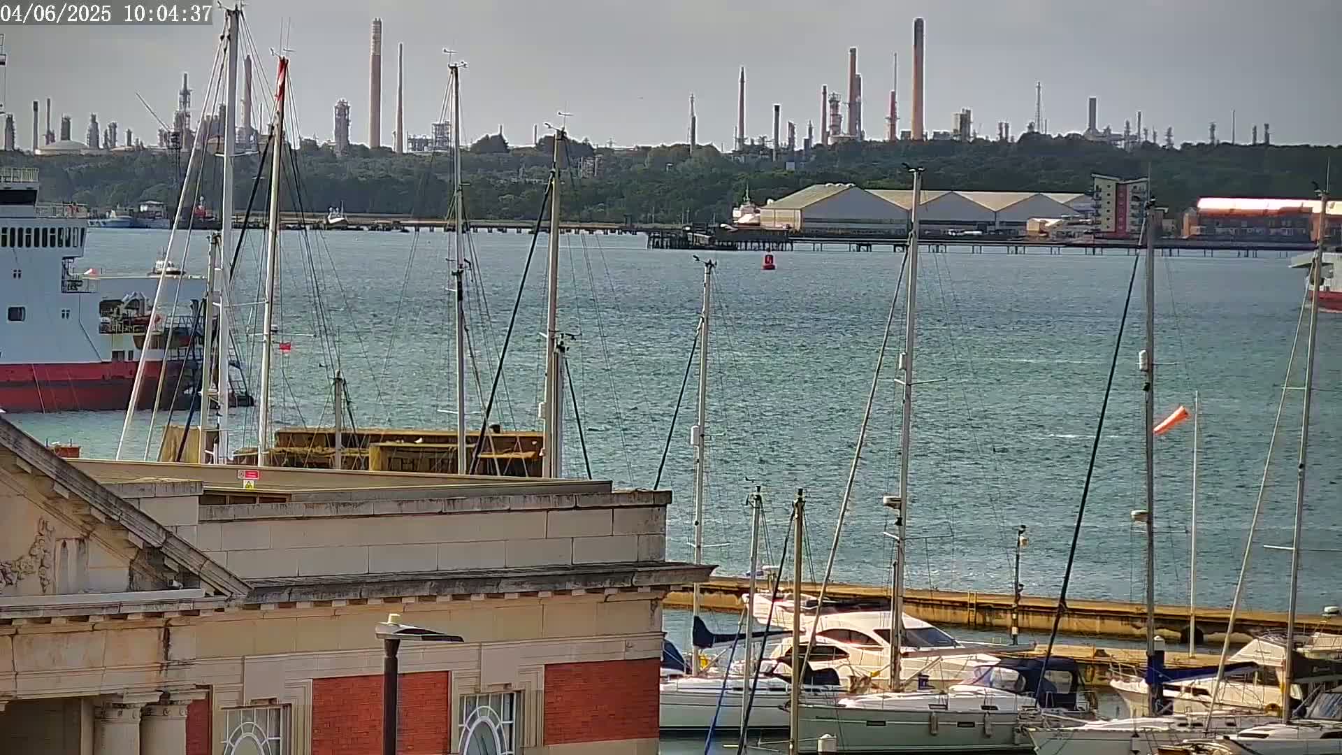 A marina filled with sailboats and motorboats is seen in the foreground, with a large cargo ship and an industrial refinery in the background on a mostly clear day.