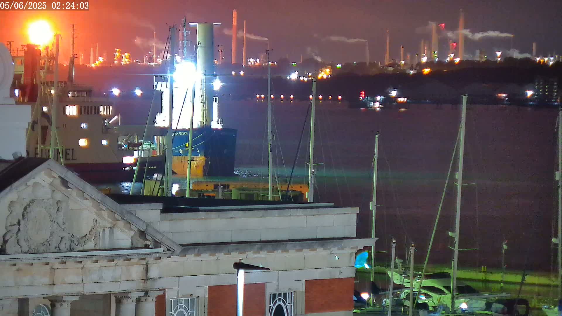 At night, a brightly lit cargo ship is docked near several sailboats, with an industrial plant glowing in the background under a dark sky.