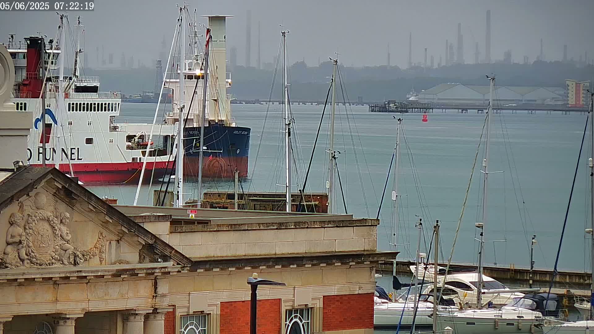 A ferry and cargo ship are moored in a harbor filled with sailboats under an overcast sky, with an industrial skyline visible in the distance.