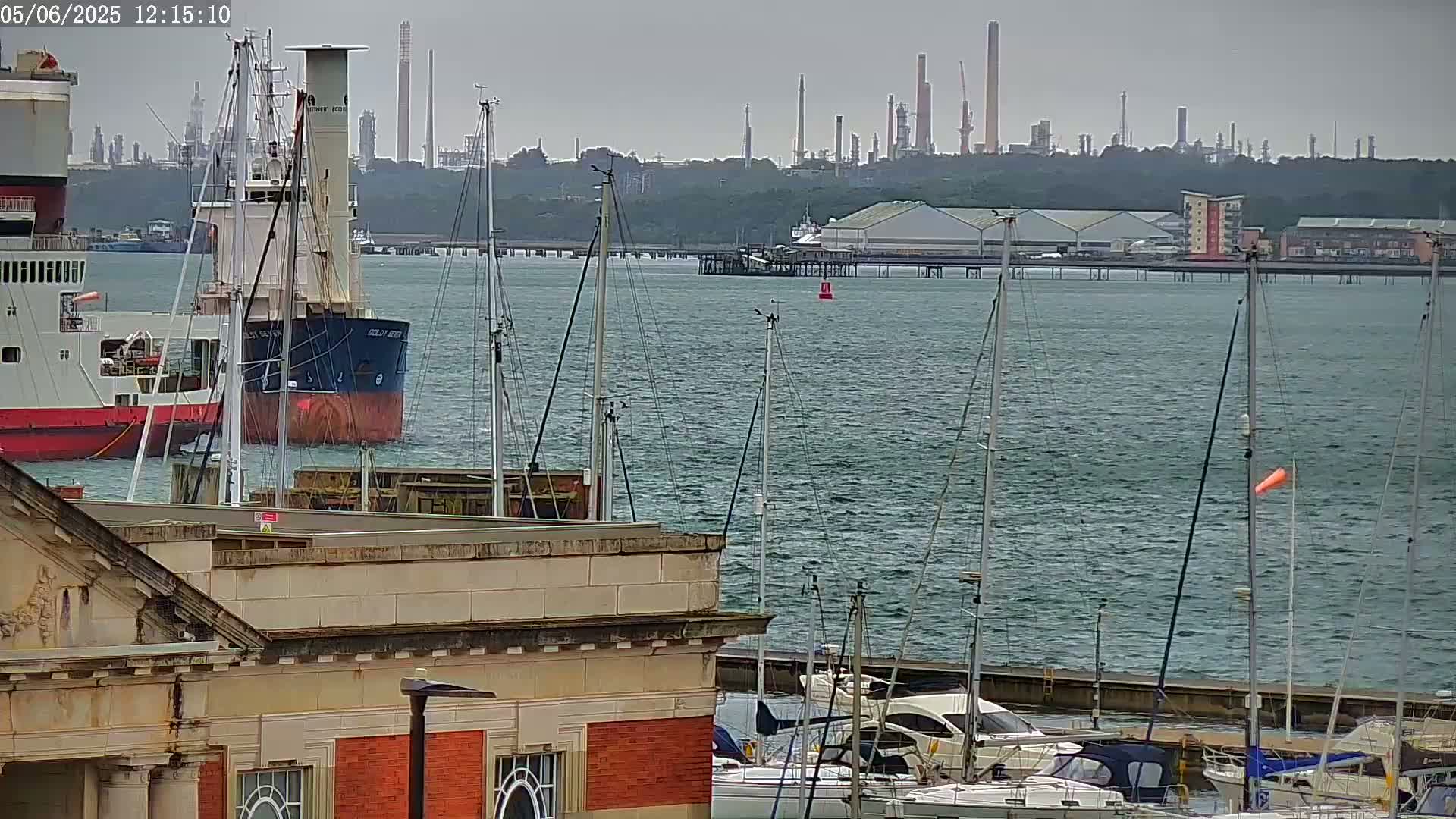A cargo ship and numerous sailboats are moored in a harbor on an overcast day, with an industrial refinery visible in the background.