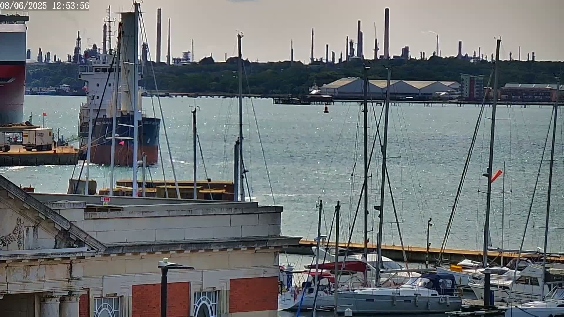 A cargo ship is moored near a marina filled with sailboats on a calm, sunny day, with an industrial refinery visible in the background.