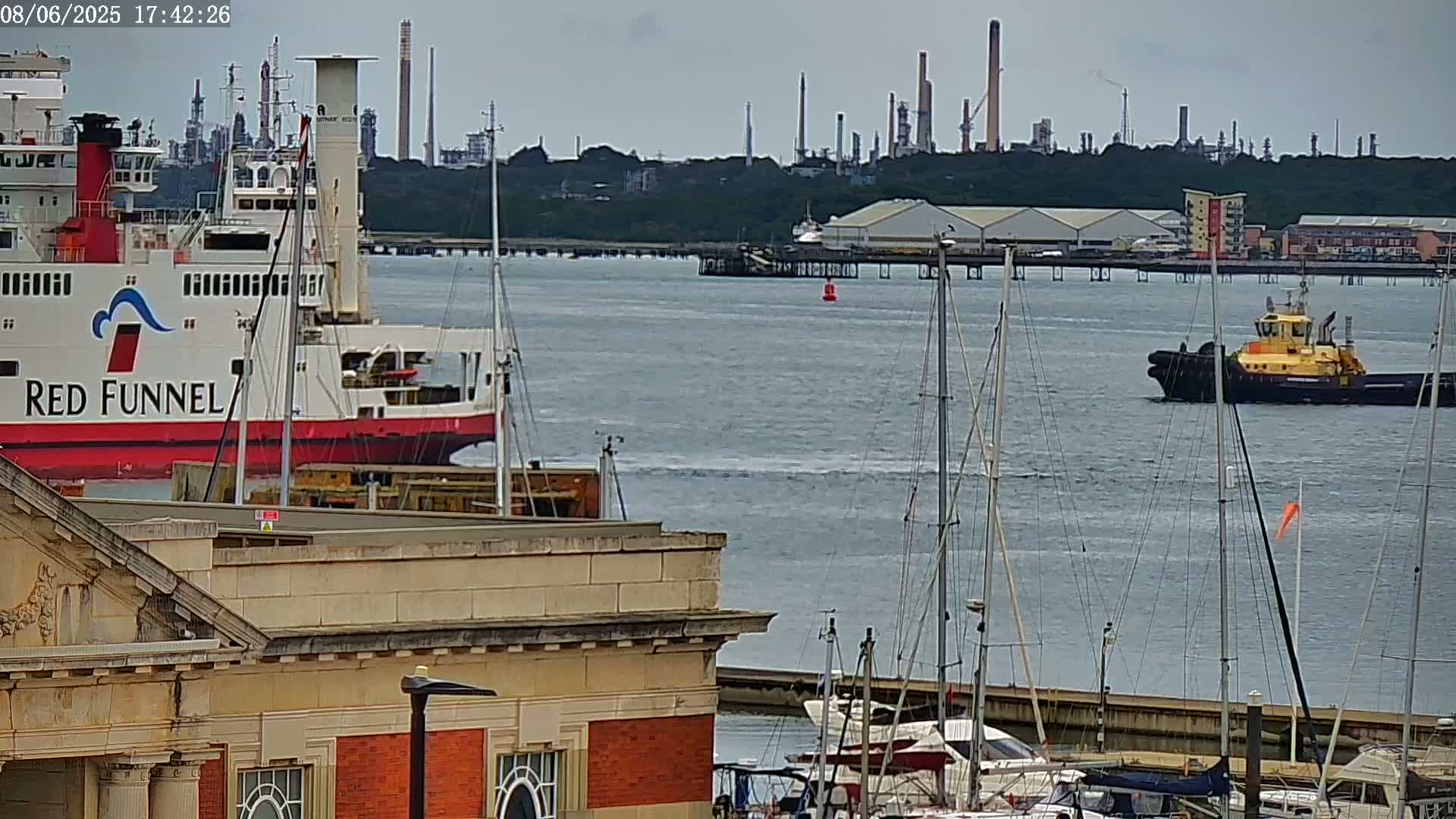 A Red Funnel ferry is docked near a marina with numerous sailboats, a tugboat is in the water further out, and an industrial complex is visible in the distance under an overcast sky.