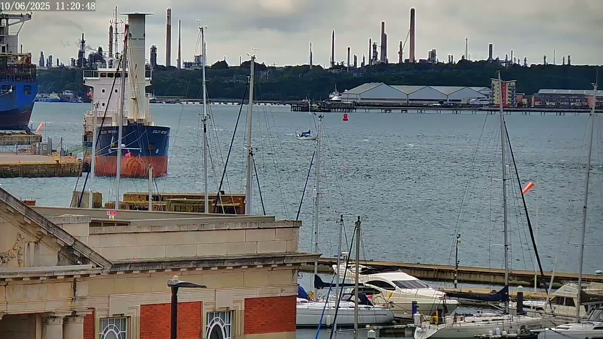 A cargo ship is moored near a harbor filled with sailboats and motorboats, with an industrial plant visible in the background under an overcast sky.