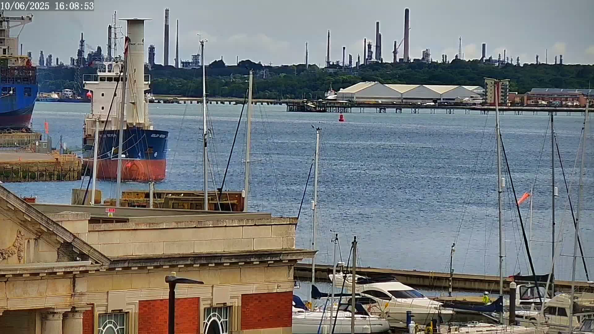 A cargo ship is docked near a marina with several sailboats and motorboats, all situated in front of an industrial refinery on a partly cloudy day.