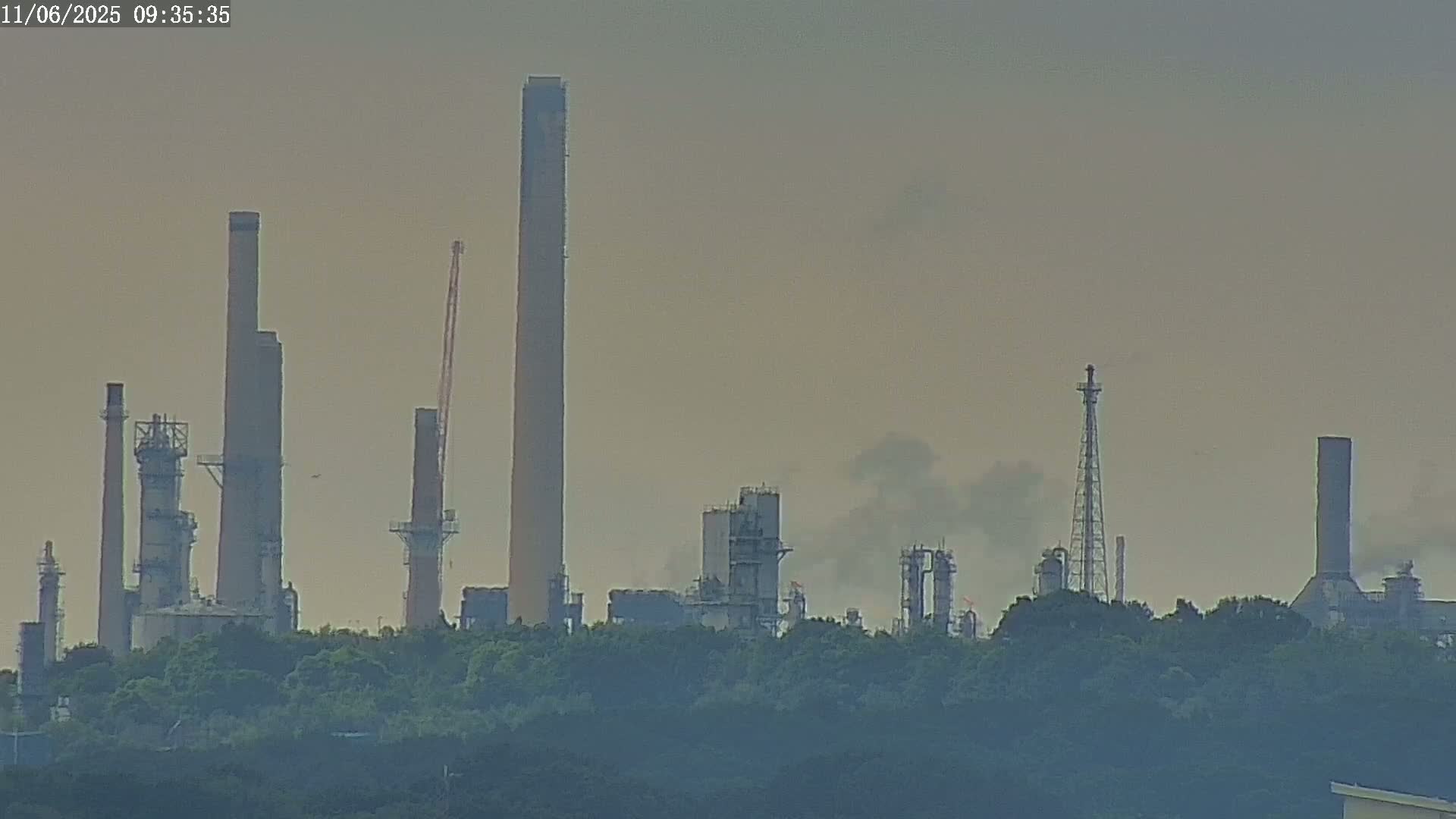 A hazy view of an industrial complex with numerous tall structures and smokestacks, partially obscured by trees in the foreground.