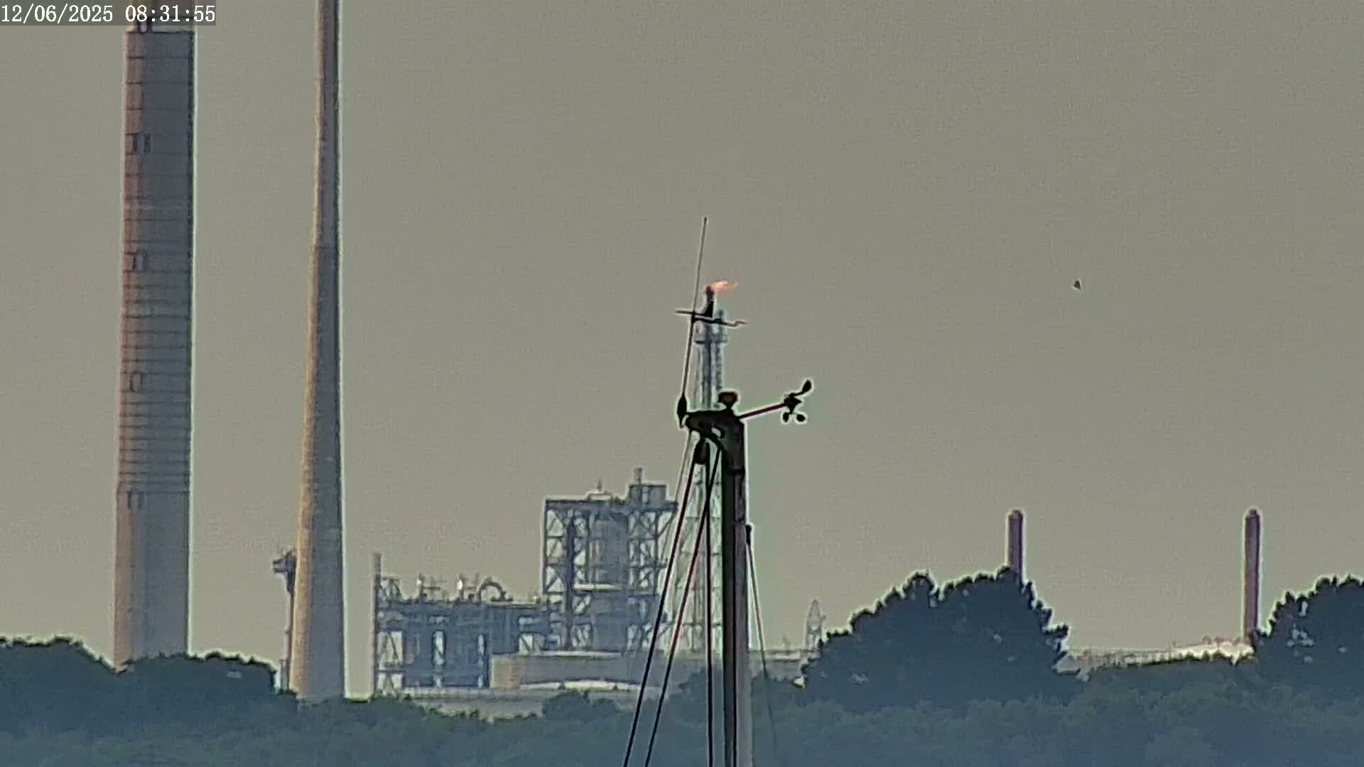 A weather vane atop a mast sits in front of an industrial plant with tall smokestacks under hazy skies.