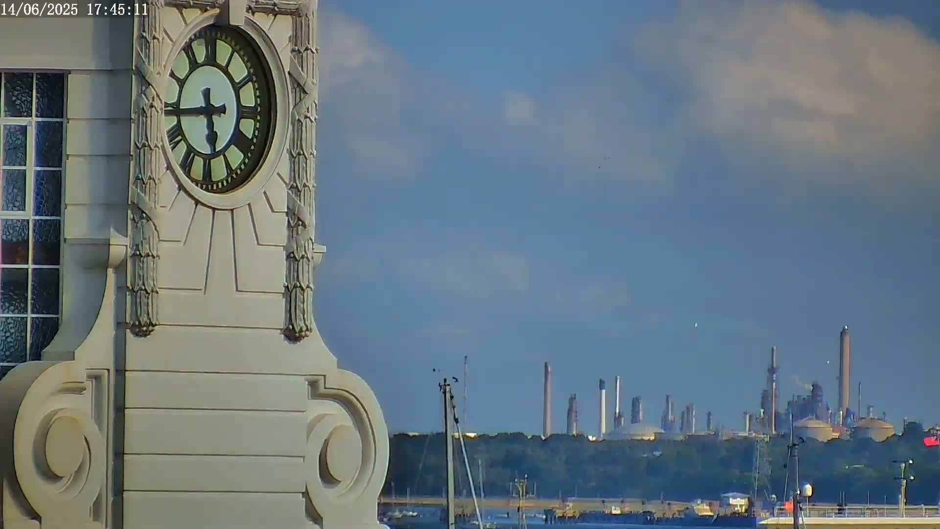 A portion of an ornate clock tower is shown in the foreground, with an industrial complex visible in the hazy distance under a partly cloudy sky.